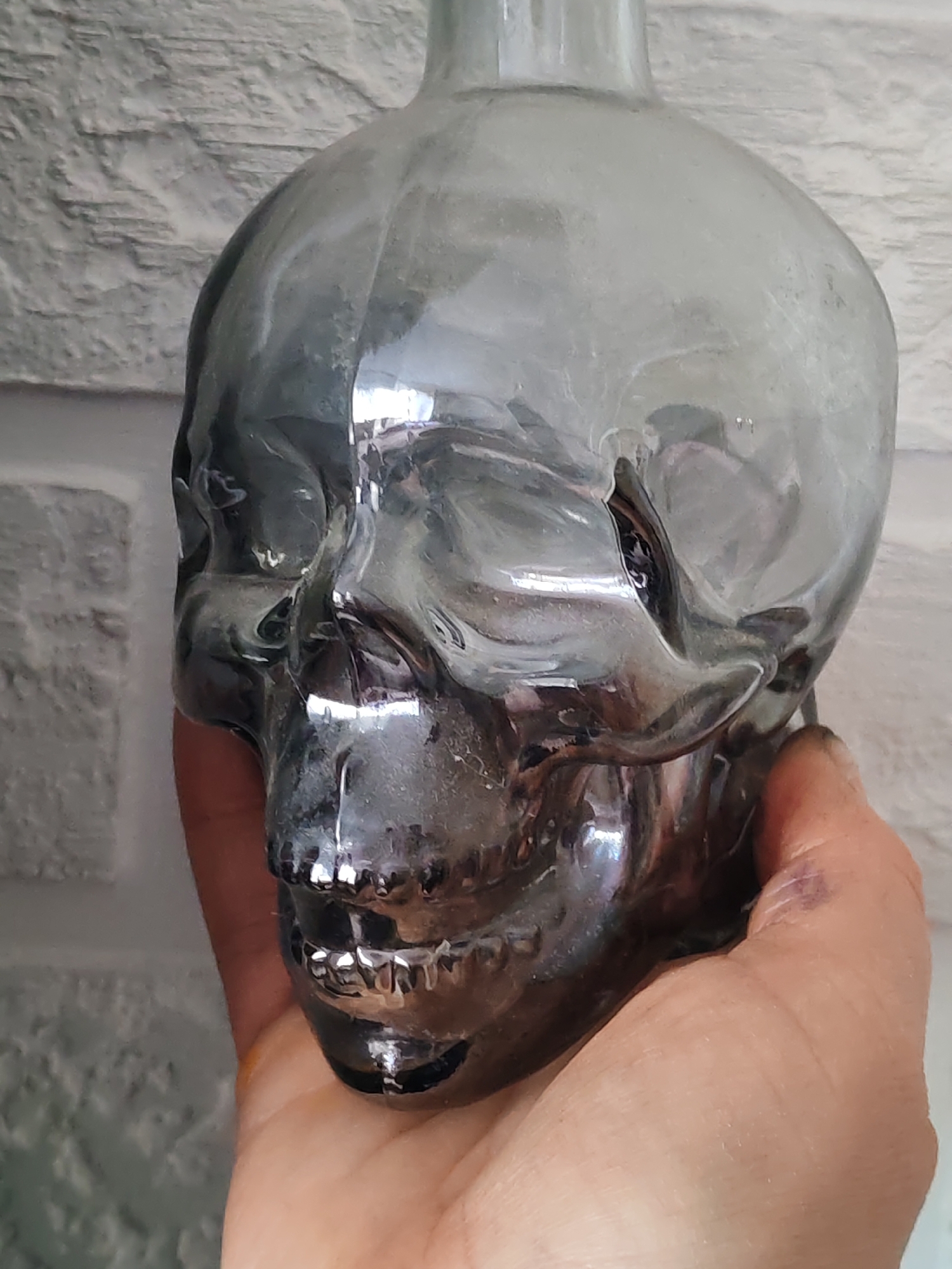 A clear, glass skull-shaped bottle is held in a person's hand, against a white brick-patterned wall. The location and city are unknown.