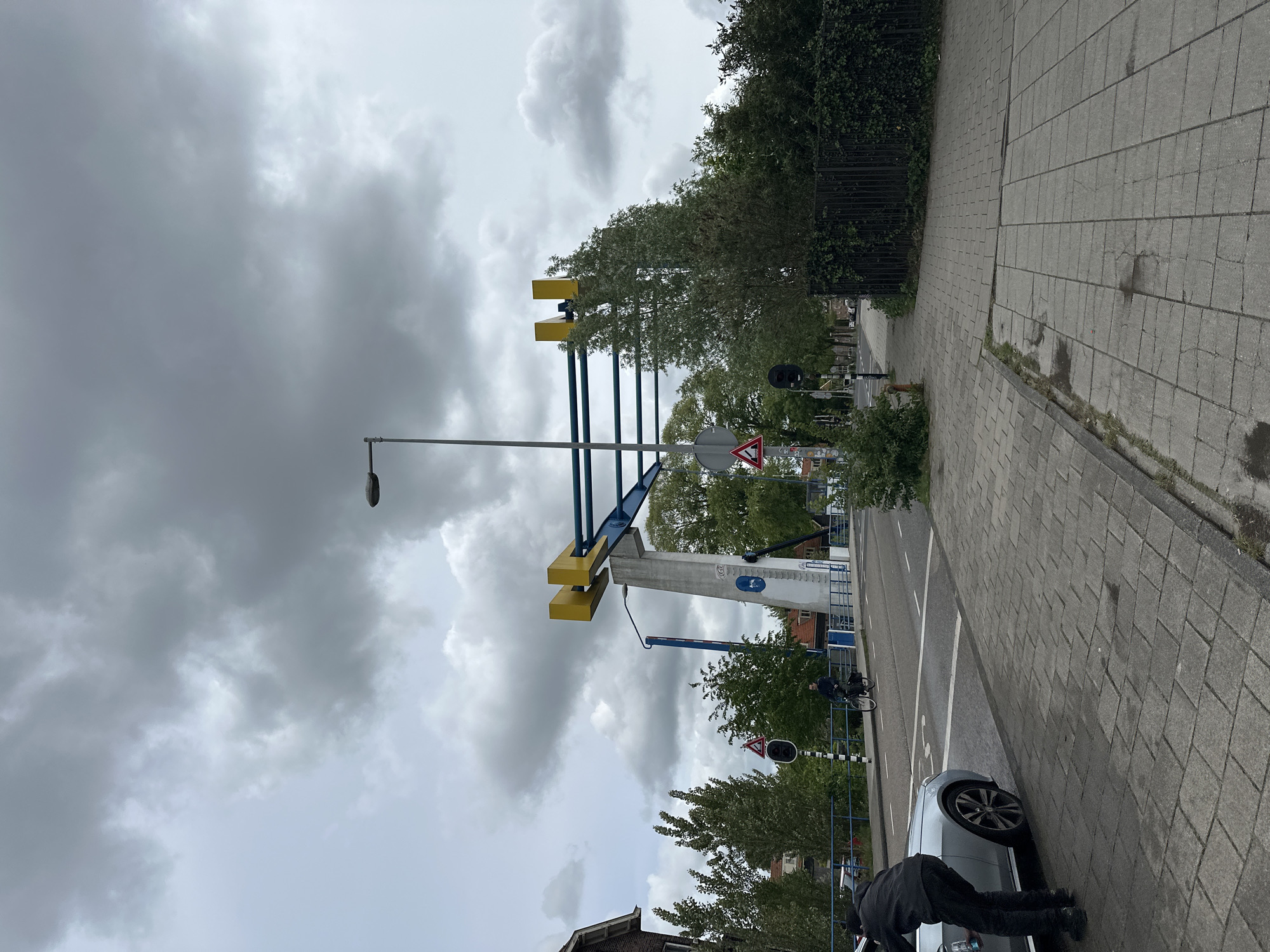 The image depicts a street scene in Amsterdam, Netherlands. A large, yellow and blue metal structure, likely a bridge support or gate, dominates the view. A street lamp is positioned near the structure, and road signs are visible. A grey car is parked on the side of the street with a person near it, appearing to interact with the car. A person on a bicycle is seen riding on the street. The street is paved with grey bricks and bordered by trees and vegetation. The sky is overcast with heavy clouds.