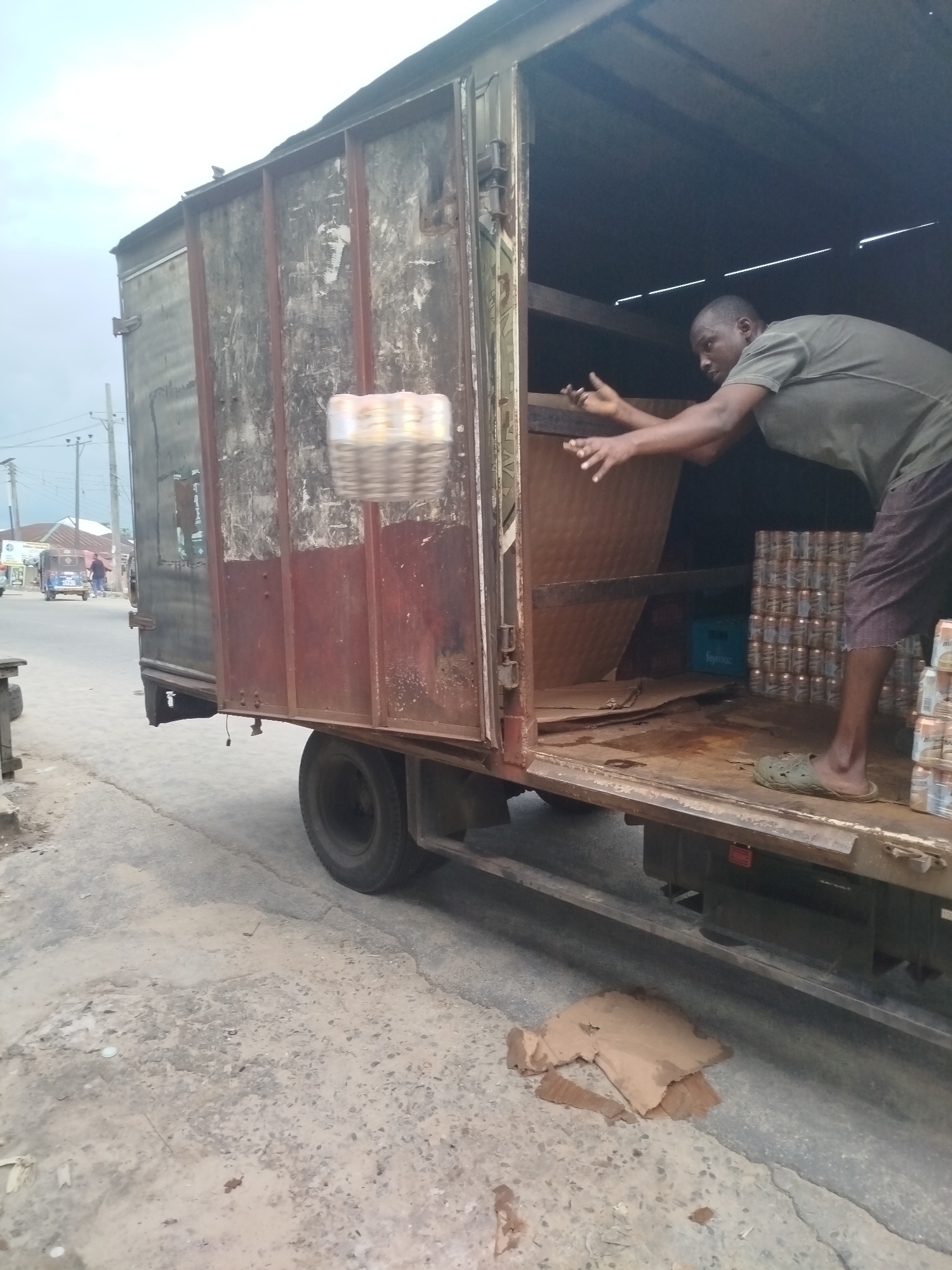 A man is unloading cans from a truck in Yenegoa, Nigeria. The truck is open at the back, revealing stacked cans inside and on the ground. The man, wearing a green t-shirt and brown shorts, is reaching into the truck with his arms extended, seemingly to grab more cans. He is also wearing green slippers. The scene appears to be outdoors, with a cloudy sky overhead. The truck is parked on a paved road. In the background, there are buildings, some power lines, and a blue vehicle that looks like a tuk-tuk. The overall impression is of a typical day in a busy area, likely a market or loading dock. The truck itself looks weathered and well-used, suggesting it's a work vehicle. The ground is concrete with some debris, including pieces of cardboard. There is no visible text in the image.
