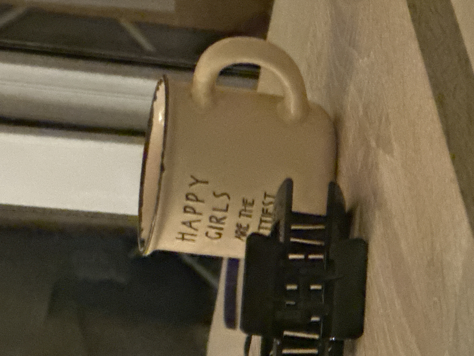 A close-up shot depicts a mug with the inscription "HAPPY GIRLS ARE THE . . . TIEST." A black hair clip is positioned in front of the mug. The background includes a window and a light-colored surface.