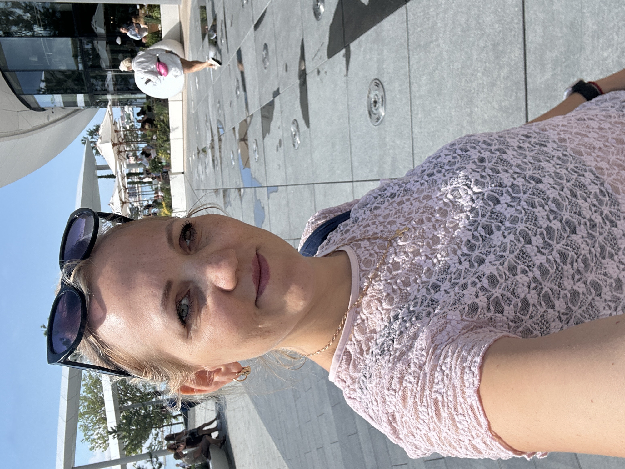 A woman in a light pink lace top stands in a plaza with a wet, tiled ground, possibly a dry fountain area. She is facing the camera and taking a selfie, with sunglasses on top of her head and a necklace visible. In the background, people are seated at outdoor tables under umbrellas, and a woman in a white shirt and pink fanny pack walks away from the camera. The location is the embankment in Gelendzhik, Russia, near the "Azure" shopping and entertainment center. The sky is clear and blue.