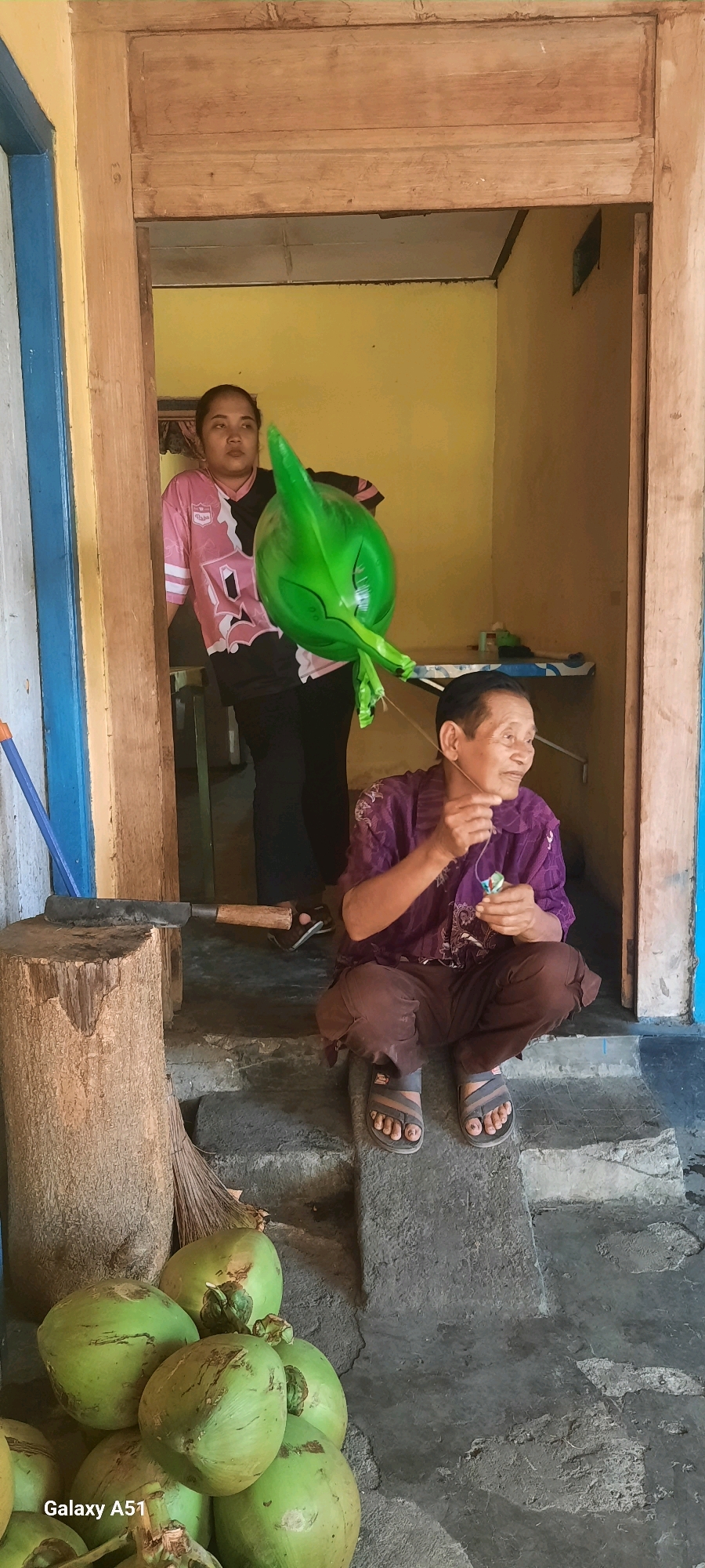 In Selopuro, Indonesia, two individuals are present. In the background, a woman stands holding a large, green, alien-shaped balloon. In the foreground, an older person is crouched on steps, holding a string with a small object attached. Several green coconuts are piled on the ground to the left of the frame, next to a wooden chopping block with a large cleaver. The environment appears to be an outdoor setting adjacent to a building with yellow walls and wooden doorframes.