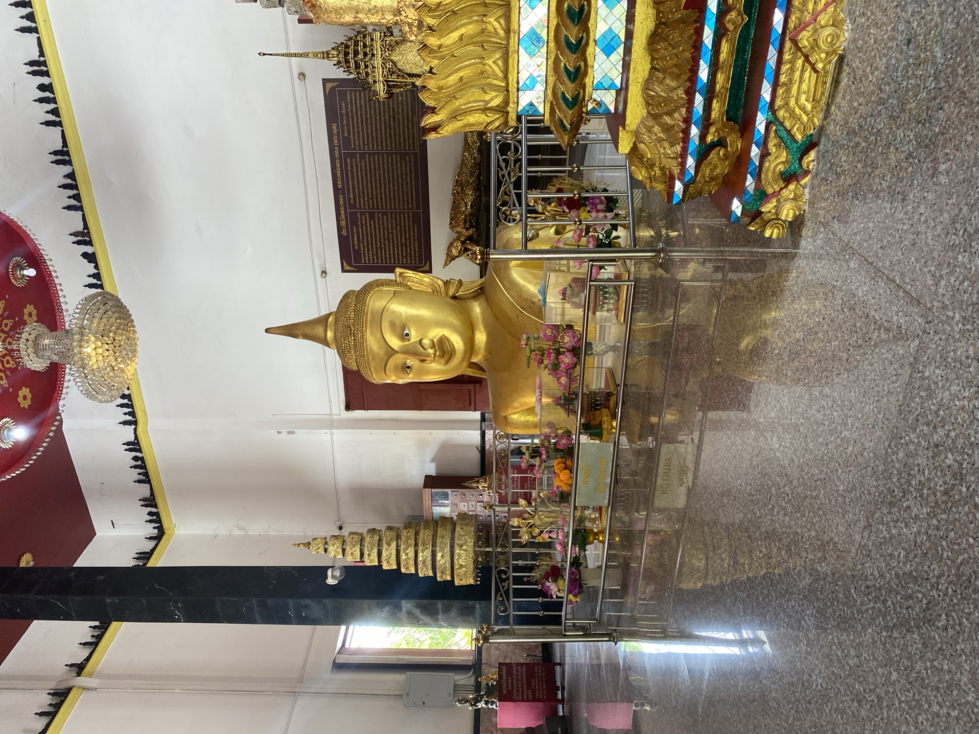 This image captures the interior of a temple, likely in Thep Krasatti, Thailand, focusing on a large, golden Buddha statue. The central subject is the head and upper torso of a Buddha, cast in bright gold, exhibiting a serene expression. The statue is positioned behind a metal barrier, which is decorated with clusters of artificial pink and purple flowers.
To the left of the main Buddha statue, a smaller, golden multi-tiered spire or stupa-like structure is visible, resting on a dark base. The temple interior features white walls, with a section of the wall and ceiling on the far left painted red, from which an ornate crystal chandelier hangs. The walls are edged with intricate black decorative patterns. The floor is made of polished granite, reflecting the golden statue and the interior lighting, which suggests a well-lit environment.
Behind the golden Buddha, a dark brown sign on the white wall displays text in Thai, which translates to "History of Wat Phra Thong - Luang Pho Phra Thong (The Emerged Buddha)" and "Legends, Tales - Stories," providing historical or contextual information about the deity. A yellow sign attached to the metal barrier explicitly warns "ห้ามเข้า" (No Entry) in Thai and "NO ENTRY" in English, indicating a restricted access area around the sacred image. The scene portrays a reverent and culturally significant setting.
