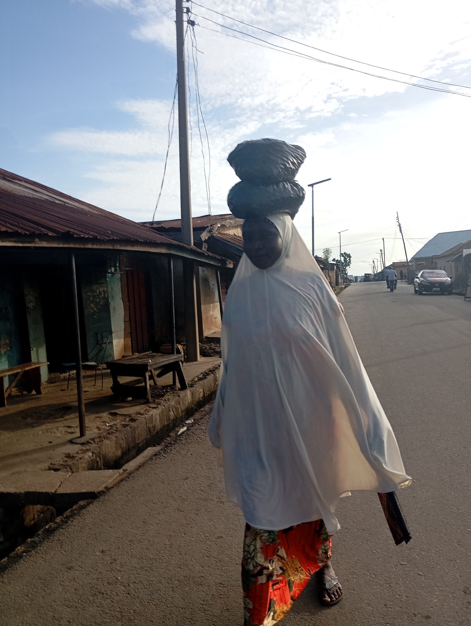 A full-body shot captures a person carrying a load of black bags balanced on their head. The individual is walking along a paved street in Shendam, Nigeria, towards the right side of the frame. They are covered in a flowing white garment, and beneath it, a vibrantly patterned skirt is visible. The person is wearing dark sandals. To the left of the street, a weathered building with a corrugated iron roof stands. The building has an open, covered porch with wooden benches and tables. The background reveals more buildings, a parked car, and a motorcycle with a rider. Overhead, utility poles with wires stretch across the sky, which is partly cloudy and bright. The scene appears to be set during daylight hours.