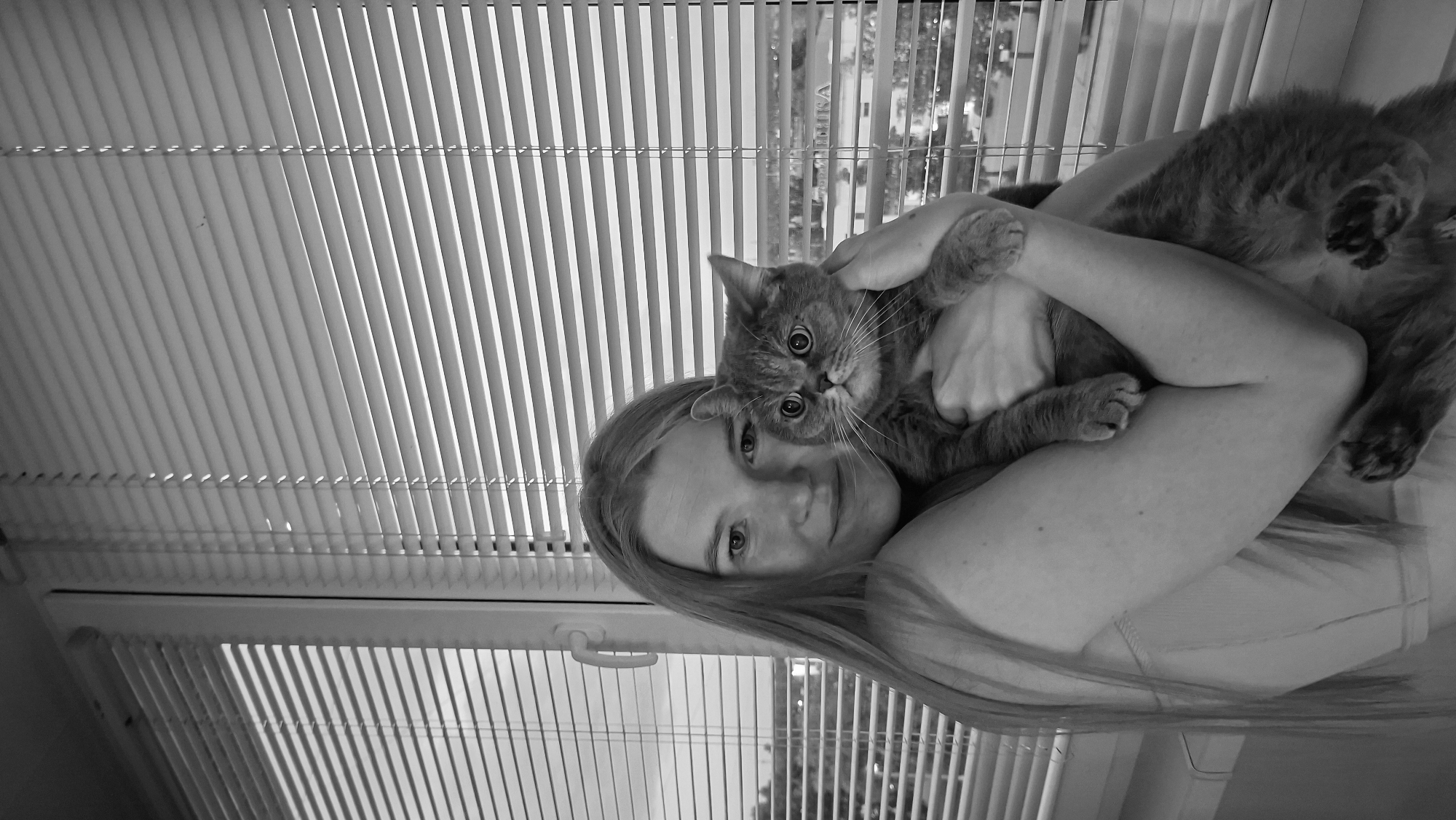 A grayscale, medium shot selfie captures a young woman with long, light-colored hair holding a gray cat. The woman is positioned on the left side of the frame, her head tilted slightly towards the camera and her gaze directed forward. She is wearing a light-colored top. The cat is held securely in her arms, its head and upper body visible. The cat has large, round eyes and appears to be looking directly at the camera. The background consists of a window with horizontal blinds, through which a blurry cityscape is visible. Text can be seen on a building in the distance, appearing to be in Cyrillic script. The scene is indoors, likely in an apartment, given the window and blinds. The lighting suggests it is daytime. No specific landmarks or distinguishing features of Chaikovsky, Russia are identifiable from the image.