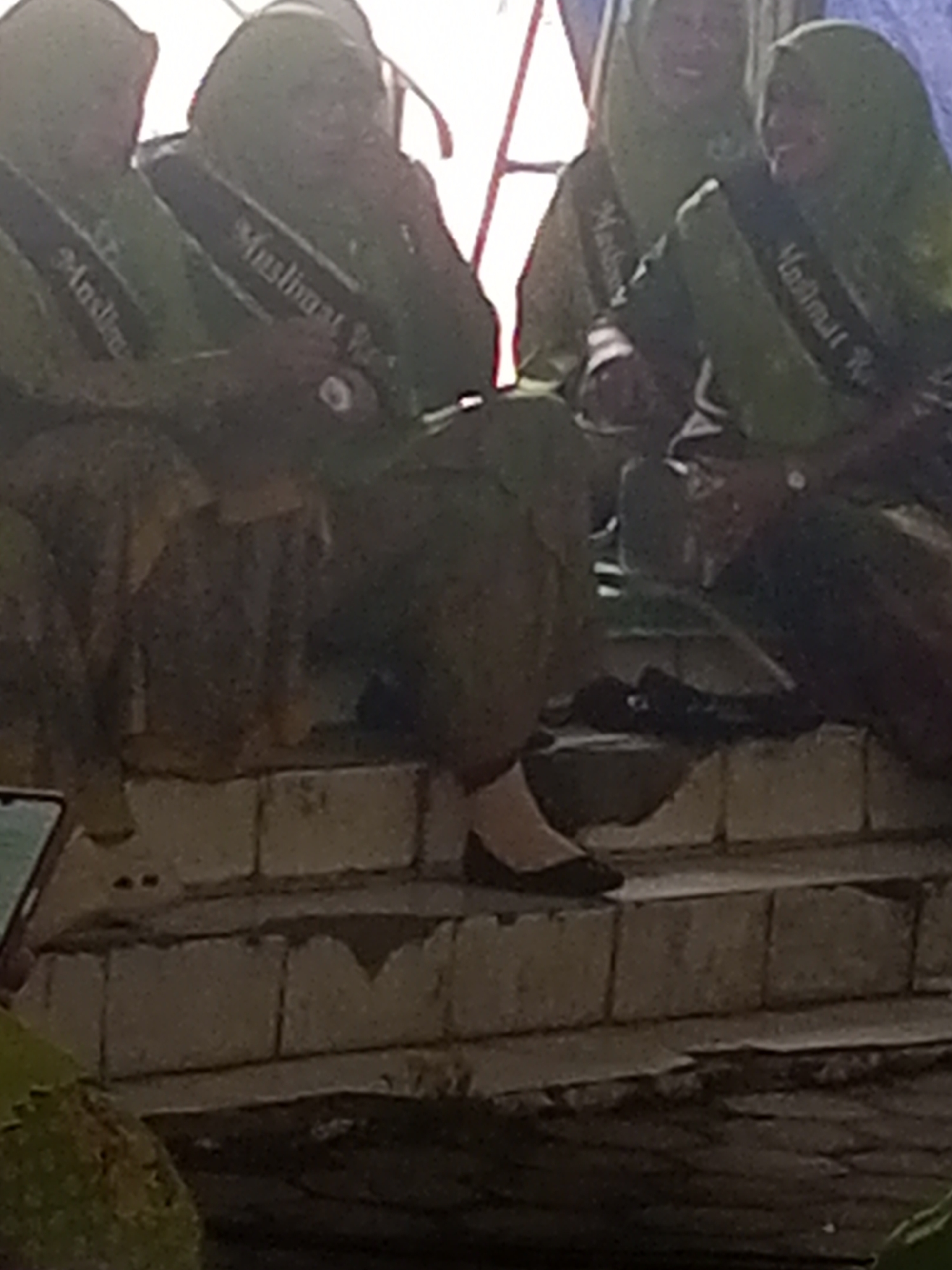 This image depicts a group of individuals, likely young women, dressed in green attire and hijabs, adorned with sashes. The sashes appear to have text, possibly indicating an event or organization. They are seated on a tiered structure resembling steps, constructed from concrete blocks. The background suggests an outdoor setting, with hints of a canopy or tent structure and possibly some greenery in the foreground. The overall atmosphere seems to be one of a ceremony or gathering. The text on the sashes are partially visible but appear to read "Muslimat" and possibly "R" or "Ri" followed by another letter. The location is identified as Klaseman, Indonesia.