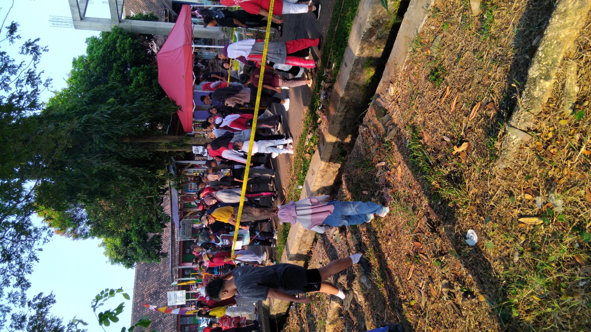The image captures a lively outdoor gathering, likely a community event or festival, taking place in the city of Posong, Indonesia. A crowd of people, including adults and children, are present, mingling and observing. Several individuals are wearing casual clothing, while some are in athletic wear, suggesting a possible sporting event or physical activity.
A yellow barrier tape is prominently featured, dividing the area and guiding the flow of people. There are tents set up, one with a vibrant red canopy, which might be serving as stalls or information booths. The background reveals buildings with tiled roofs, indicating a typical Indonesian urban or semi-urban setting. Trees with dense foliage provide shade, and the ground is covered with dry leaves, grass, and some debris, characteristic of an outdoor environment.
The scene appears to be during the daytime, with sunlight casting shadows and highlighting the various elements. The overall atmosphere seems to be one of activity and engagement, with people spread out and interacting within the designated space.