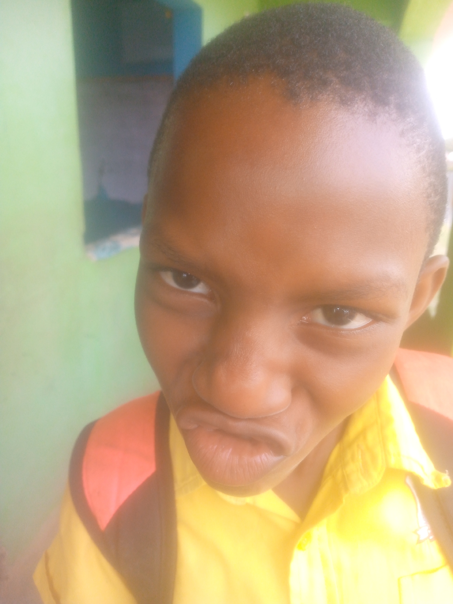 A close-up, medium-shot photograph captures a young, dark-skinned individual with a closely shaved head. The person is wearing a yellow shirt with a red and black backpack visible behind them. The background features a green wall with a partially visible blue cabinet or structure in the upper left corner. The individual's facial expression is noteworthy, with a slightly puckered mouth. The location appears to be indoors. There is no identifiable location within the city of Unknown, Unknown.