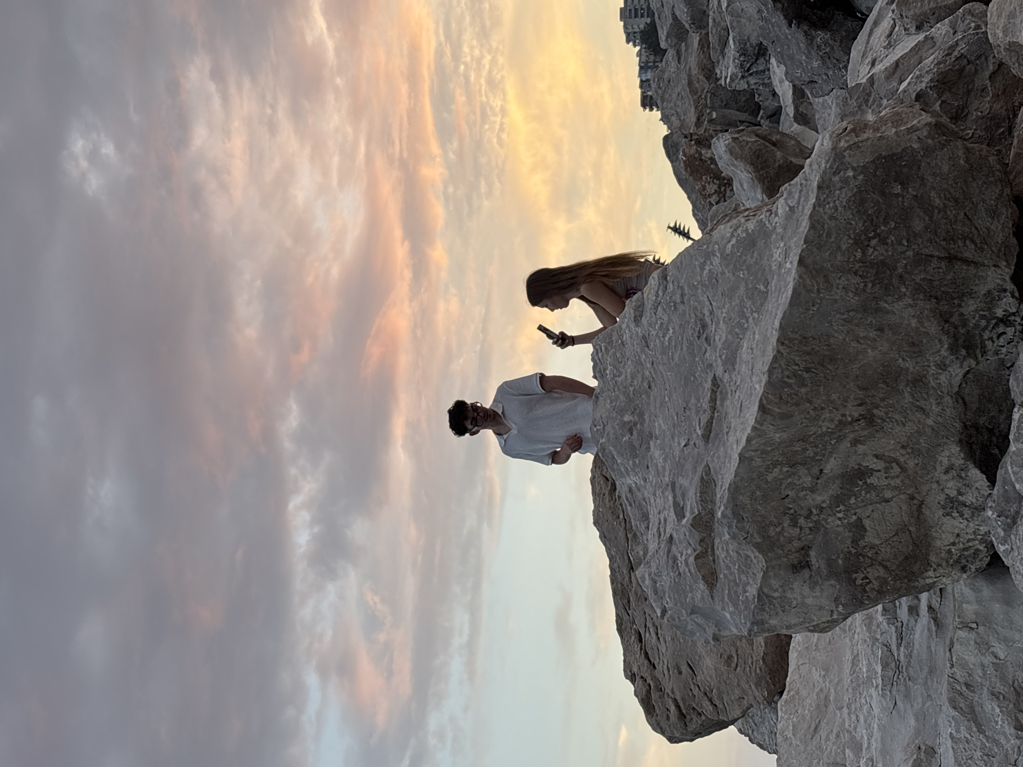 A young man wearing glasses and a white polo shirt stands on large rocks against a twilight sky. To his right, a young woman with long, light brown hair, wearing a dark top, holds a smartphone and appears to be taking a picture or recording. They are positioned on a rocky shore, possibly near the city of Budva, Montenegro, given the buildings visible in the distance on the right side of the frame. The sky is filled with soft, pastel-colored clouds, indicating either sunrise or sunset. The foreground is dominated by the rough texture and varied sizes of the large rocks.