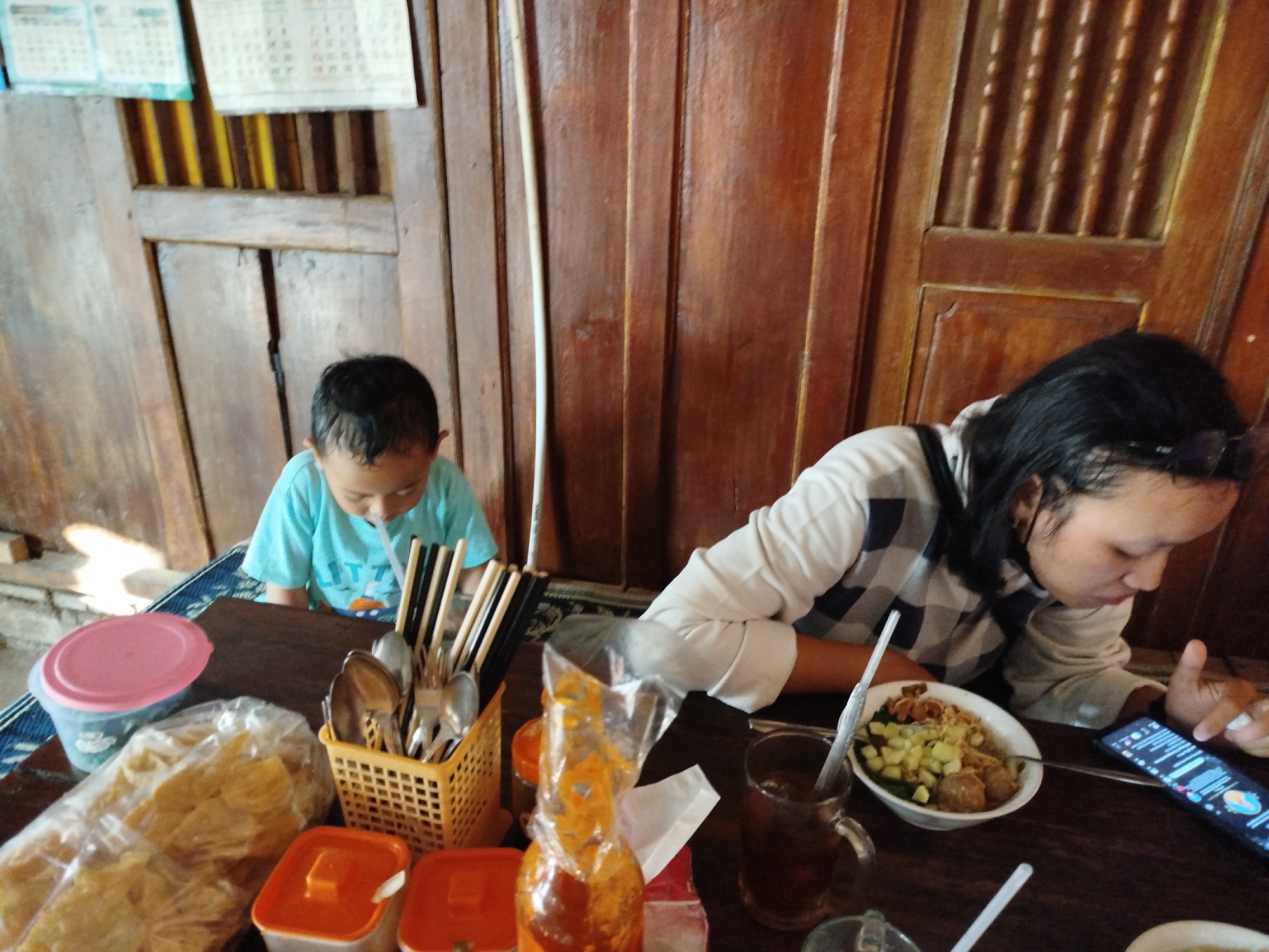 The image captures an indoor scene, likely a casual dining establishment, with a prominent wooden paneled wall and door in the background. On a dark wooden table in the foreground, a young child wearing a light blue t-shirt is seated and appears to be drinking from a straw. To the right of the child, a collection of eating utensils, including spoons and chopsticks, are held in a yellow basket. In front of the utensils is a plastic bag filled with what appears to be chips or crackers. To the left of the basket, a pink-lidded container and a blue container are visible.
Further to the right on the table, a woman is seated, wearing a checkered long-sleeved shirt and black-rimmed glasses on her head. She is looking down at a smartphone held in her right hand, and her left hand is near a bowl of food containing noodles, meatballs, and diced cucumbers, with a spoon resting in it. To the left of the food bowl is a glass of dark liquid, likely tea, with a straw. Another glass and a bottle of orange sauce are also present on the table. The overall impression is a snapshot of a mealtime interaction between a child and an adult in a simple, perhaps rural, Indonesian setting. The location is identified as Pojok, Indonesia.