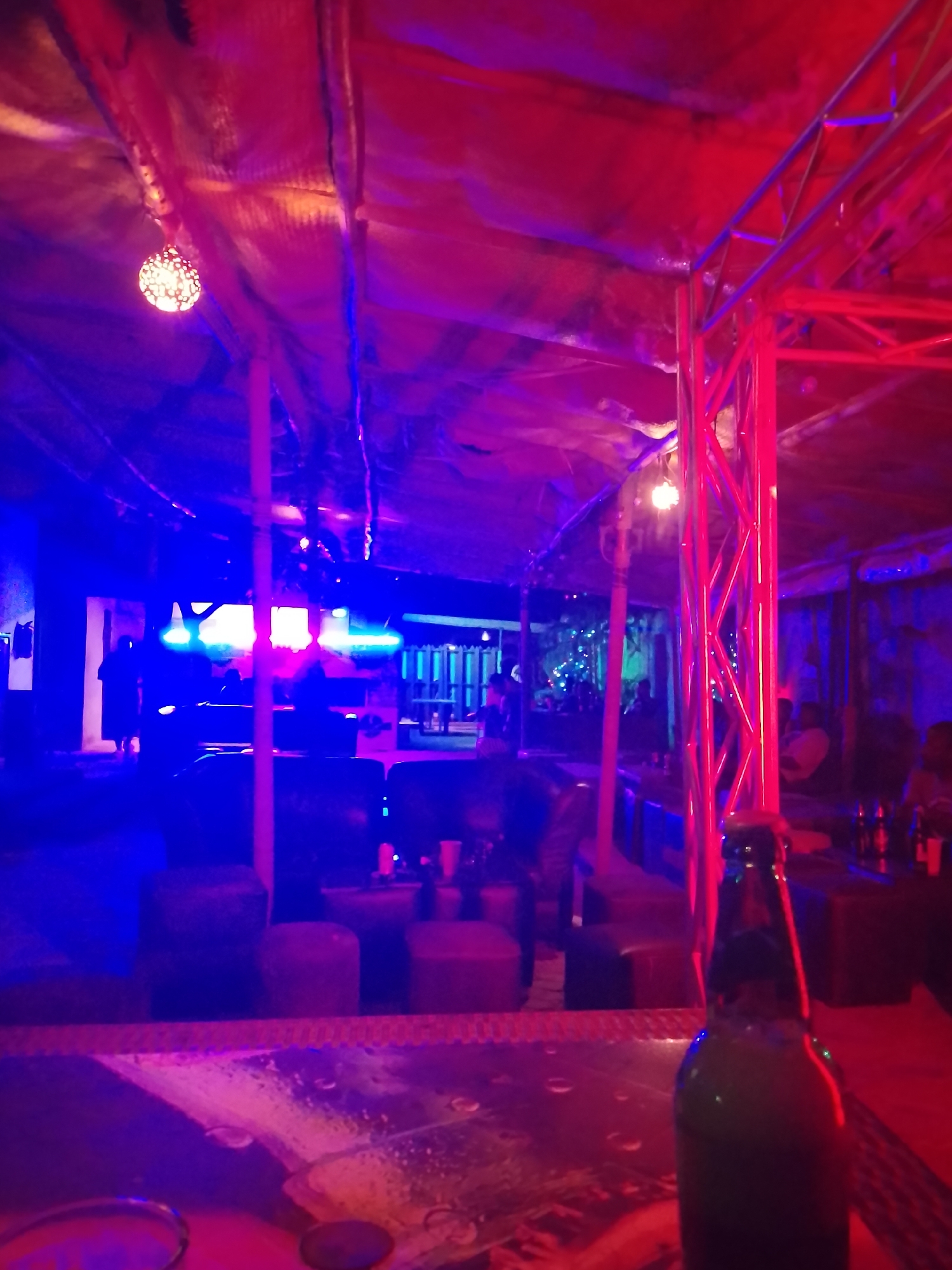 The image depicts the interior of a nightclub in an unknown location. The space is illuminated with vibrant blue and red lighting, creating a lively atmosphere. The scene features a bar counter in the foreground with a partially visible glass and a bottle. In the background, there are seated patrons, with several individuals standing near what appears to be a stage or entertainment area. The furniture includes stools, ottomans, and lounge seating. A metal support structure runs vertically through the scene, adding an industrial touch. Decorative hanging lights are also visible from the ceiling.
