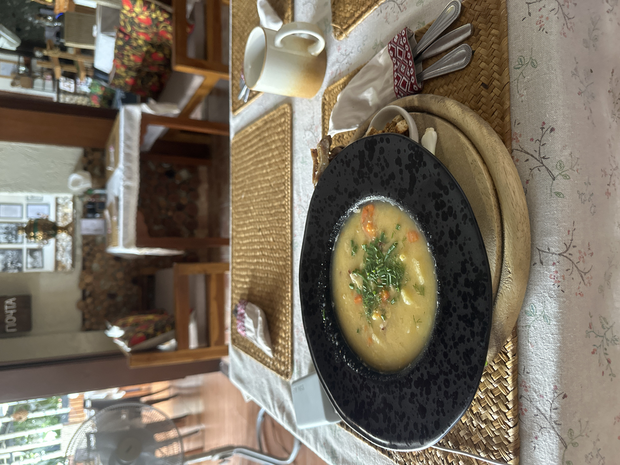 The image shows a table setting in what appears to be a restaurant or dining area. A black plate filled with soup is the central focus, garnished with fresh herbs. The plate rests on a wooden charger. Adjacent to the soup, there is a small white bowl with what appears to be bread, along with a napkin and silverware. A mug is also visible in the upper portion of the frame. The table is covered with a patterned tablecloth, with place mats underneath. In the background, there is a glimpse of the restaurant's interior, including furniture and decor. The location is suggested to be in Baan Chaweng Noi, Thailand, but there is no direct evidence of this in the image.