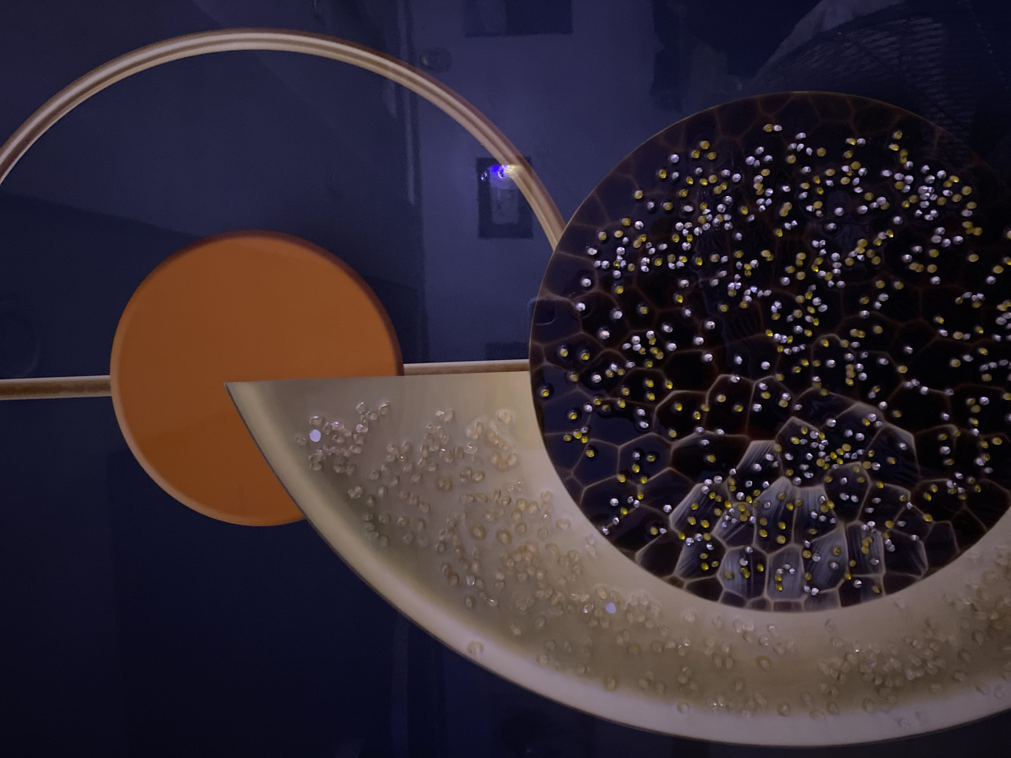 An abstract sculpture is depicted in the city of Giginyu, Nigeria. The sculpture features an orange disc at the top, a beige crescent shape with small raised bumps or dots, and a dark circular element with a honeycomb pattern filled with more bumps or dots, alternating between yellow and white. A thin, curved gold-colored metallic band partially encircles the upper right portion of the sculpture. The background is a dark navy blue, with hints of a faint purple glow and a rectangular frame visible in the distance. There are no people present in the image, and no actions or interactions are taking place.