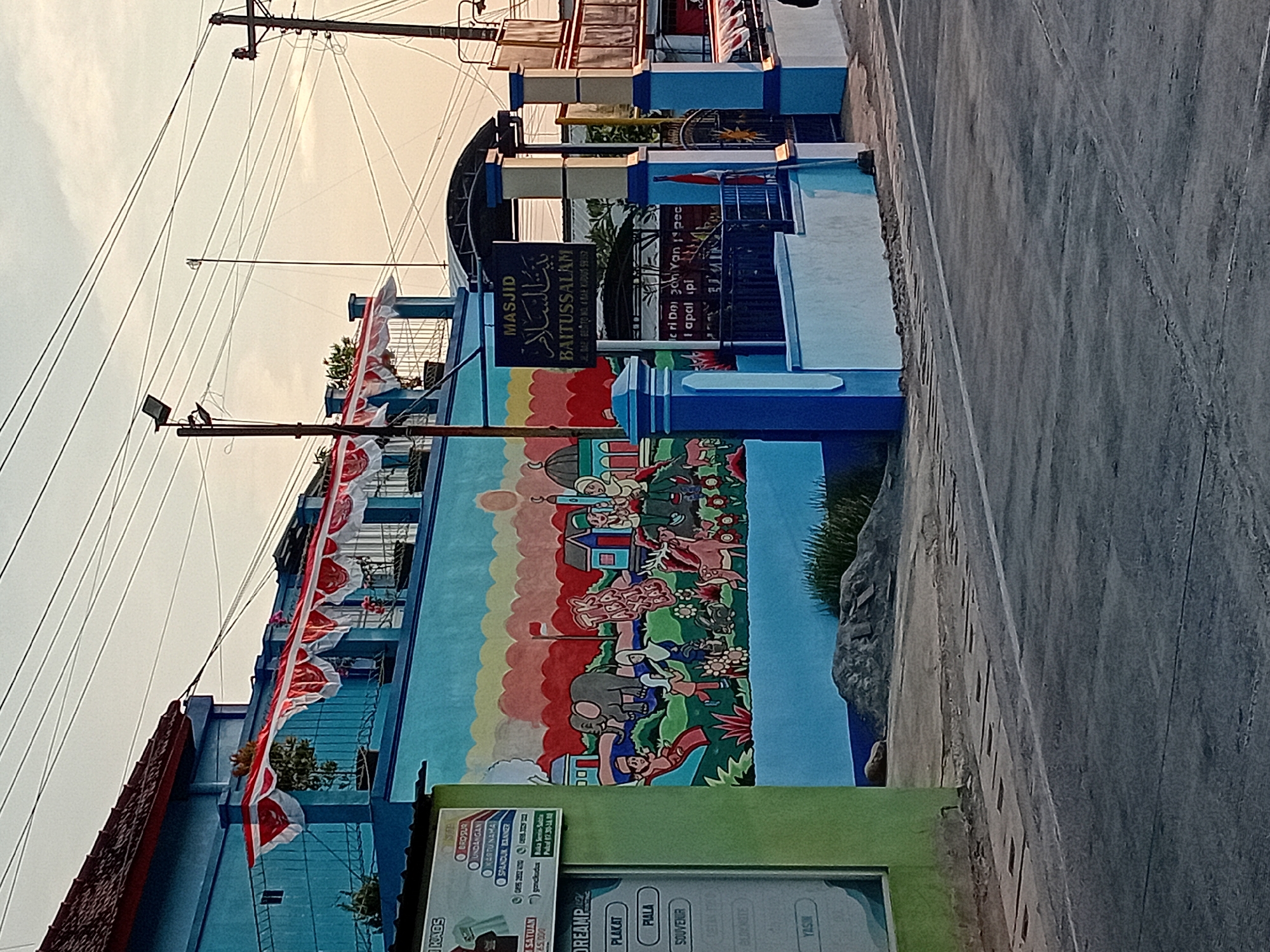 The image captures a street scene in the city of Bae, Indonesia, featuring a building with a vibrant mural on its exterior. The mural depicts a colorful landscape with stylized figures, animals like an elephant and a horse, and elements of Islamic architecture. Red and white Indonesian flags are displayed along the upper balcony of the building, suggesting a celebratory or nationalistic occasion. A sign with Arabic script and "BAITUSSALAM" in Latin letters indicates that the building is a mosque. The mosque entrance is flanked by blue and white pillars, with an iron gate. The street in front of the building is paved, with some grassy areas and construction materials visible on the side. Overhead, a network of power lines crisscrosses the sky, and utility poles are present. The lighting suggests it is either early morning or late afternoon, with soft, diffused light.