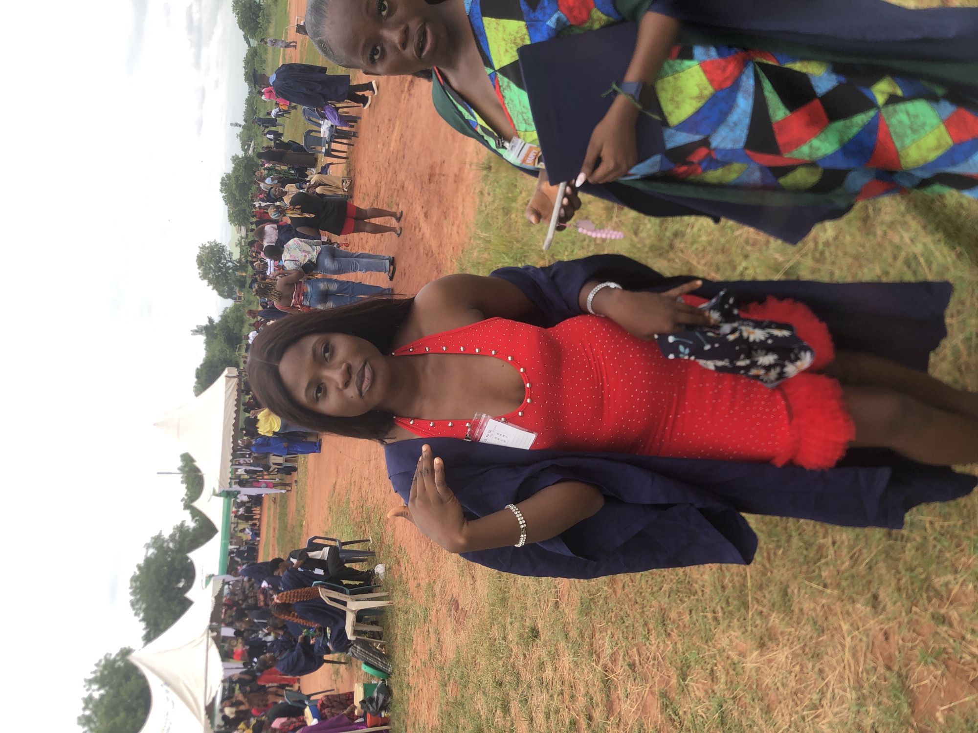 The image shows two women at an outdoor event, possibly a graduation ceremony, in Ukwu-Abwa, Nigeria. The woman in the foreground is wearing a red dress with embellishments and a dark blue graduation gown draped over her shoulders. She is also wearing a bracelet. The second woman is wearing a colorful, patterned dress and also has a graduation gown draped over her. She is holding a dark blue folder. The background shows a large gathering of people seated under white tents, with rows of chairs and trees in the distance. The ground is a mix of grass and reddish soil.