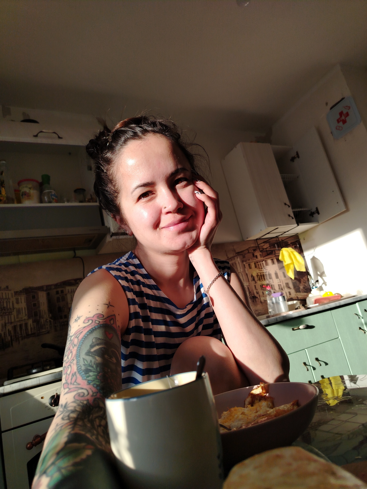 A young woman is captured in a sunlit kitchen, smiling warmly at the camera, her right cheek resting on her hand. She has dark hair tied up, a nose piercing, and is wearing a blue and white striped sleeveless top. A prominent, colorful tattoo of what appears to be a raccoon or similar animal within an ornate frame, surrounded by foliage, adorns her left arm.
The scene appears to be a casual moment, possibly during breakfast or a morning beverage. In the foreground, a white ceramic mug and a bowl containing food, likely scrambled eggs or a similar dish, are visible on a table, along with a partial view of some bread or pastry.
The kitchen features overhead white cabinets and lower cabinets painted in a light green hue. A range hood and stovetop are present on the left side of the frame, with shelves above holding various jars and bottles. The backsplash behind the counter showcases a decorative print of an old-world city scene, possibly Venice. A yellow cloth is draped near the sink area, and a light-blue rectangular object with a red cross symbol is mounted on the wall. Strong, warm sunlight streams into the room from the right, casting highlights and shadows, indicative of either early morning or late afternoon. The woman appears relaxed and content in this domestic setting in Noviy, Russia.