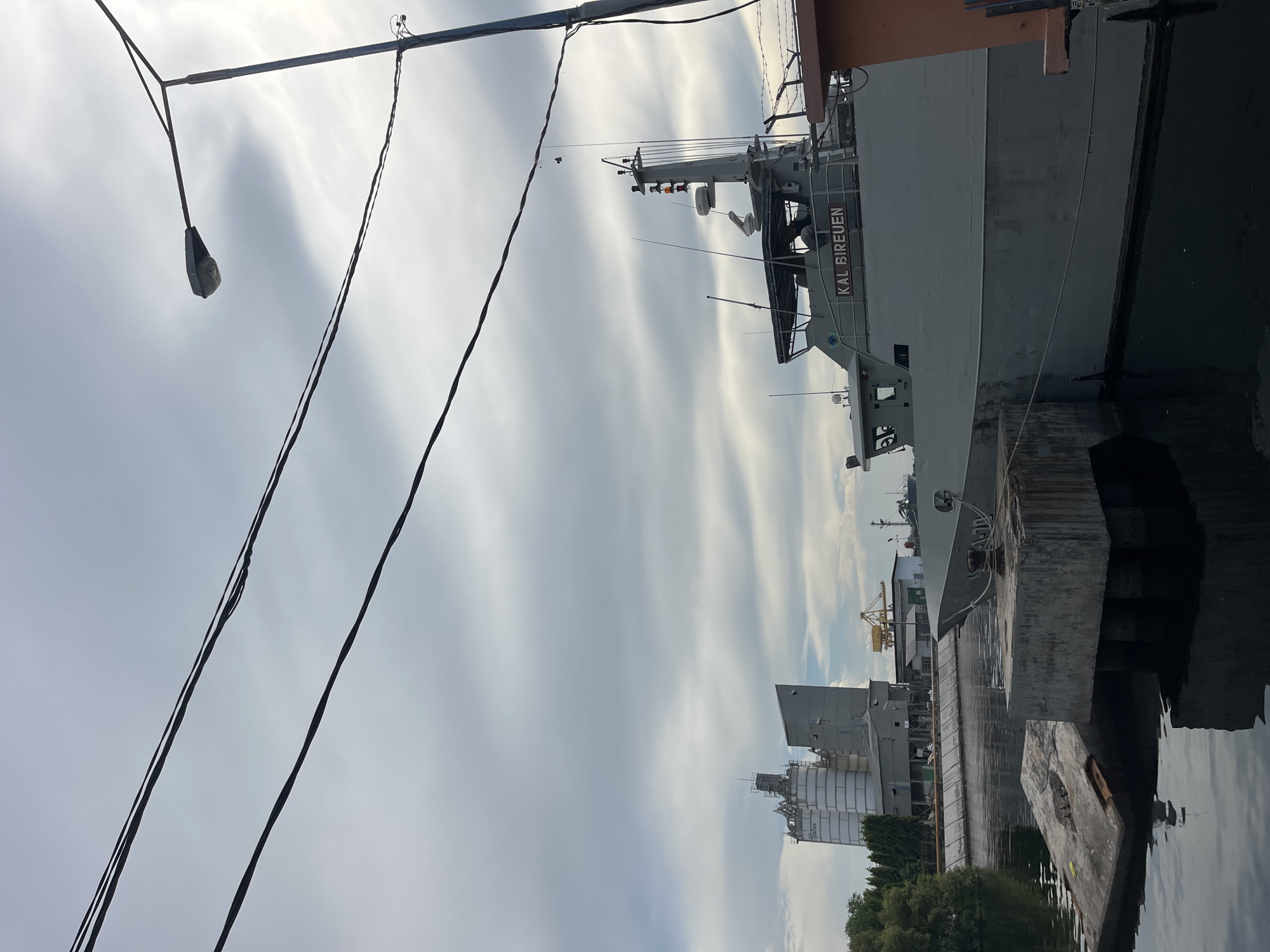The image shows a grey military ship docked at a concrete pier. The name "KAL BIREUEN" is visible on the ship's hull. To the left of the ship are industrial buildings, including large silos. A street light and several power lines are visible in the foreground. The sky is overcast with striated clouds. The scene is potentially located in Tambonbaroh, Indonesia, as implied by the ship's name, though further location specifics are unclear. Water is visible in the lower right corner of the image.