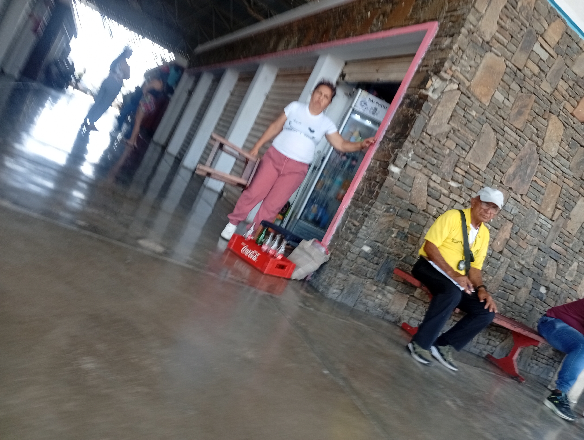 In Juan Griego, Venezuela, a woman wearing a white t-shirt and pink pants stands near a refrigerator, with a red Coca-Cola crate filled with bottles at her feet. To the right, a man in a yellow shirt and black pants sits on a red bench, holding a piece of paper, while another person in a maroon shirt and blue jeans is seated to his right on the same bench. The scene is outdoors, with a stone-tiled wall and a concrete floor.