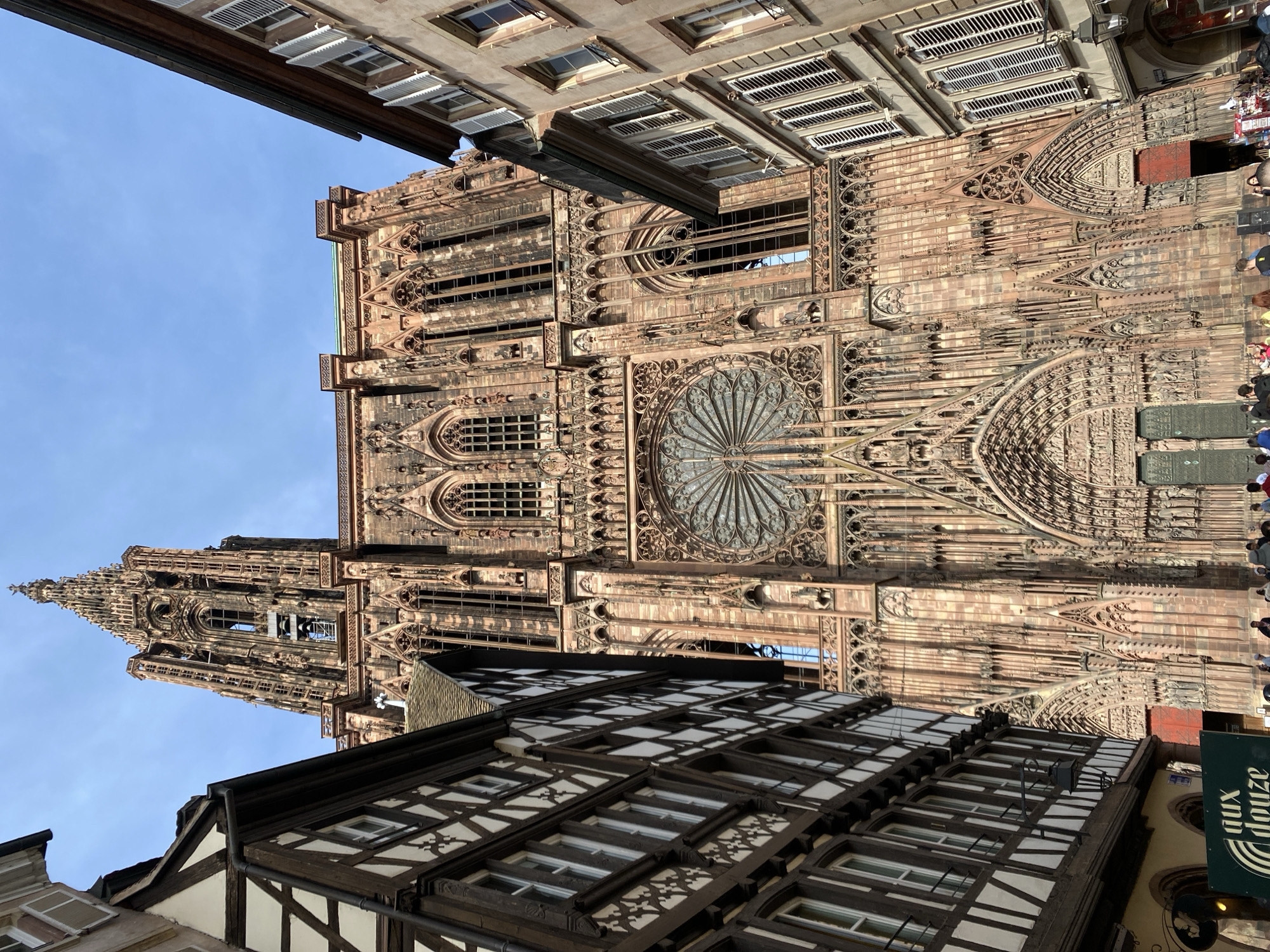 The rotated image captures the majestic Notre-Dame de Strasbourg Cathedral under a clear, bright blue sky. The cathedral's ornate Gothic facade, constructed from reddish-brown sandstone, dominates the scene, featuring intricate carvings, numerous pointed arches, and a prominent rose window with delicate tracery at its center. To the left, a tall, slender spire rises, adorned with architectural details and reaching high into the sky.
In the foreground and to the right, traditional Alsatian half-timbered buildings provide a stark contrast to the cathedral's grandeur. One such building in the immediate foreground features dark timber frames against white walls, with multiple windows. A green sign is partially visible at the bottom right corner, displaying the text "aux douze," likely indicating a business name such as a restaurant or shop.
A few indistinct human figures can be seen near the base of the cathedral, suggesting pedestrian activity in the area. The lighting indicates it is daytime, with clear, sunny weather and no visible clouds. The overall impression is one of historical beauty and architectural magnificence in the heart of Strasbourg, France.