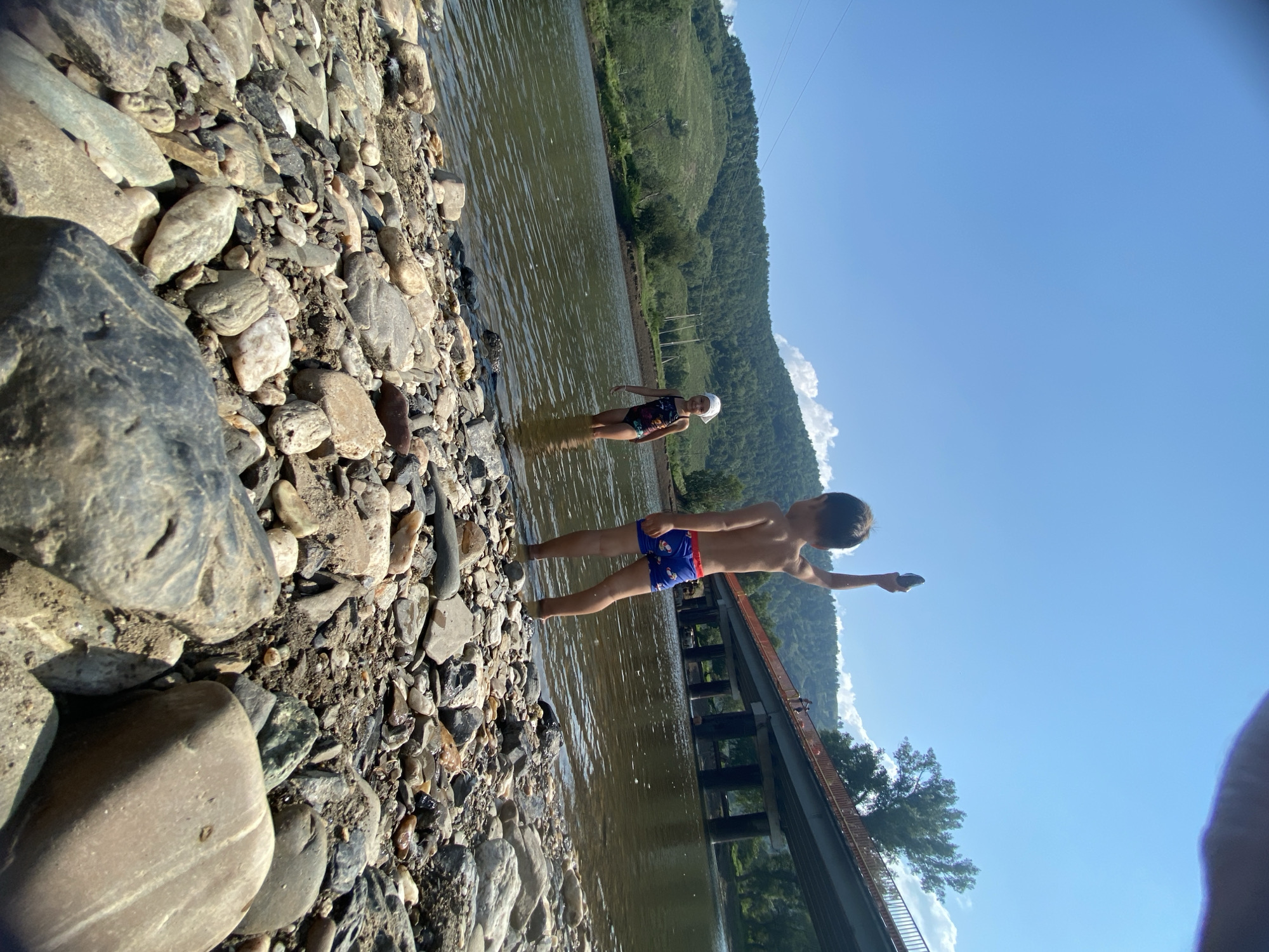 In the city of Maloarslangulovo, Russia, on a sunny day, two children are playing in the shallow water of a river. The foreground is dominated by a rocky shore, with smooth, varied-sized stones filling the frame. In the middle ground, a young boy, shirtless and wearing blue swim trunks with a red waistband, stands in the water, holding a dark-colored rock raised above his head. To his left, a young girl in a patterned swimsuit and a white headscarf stands in the river, facing the boy. The river is calm with ripples on the surface. In the background, a verdant, tree-covered hill rises steeply from the opposite bank. A large concrete bridge with an orange railing spans the river, its dark supporting pillars visible in the water. The sky is a clear, bright blue with a few wispy white clouds on the horizon.