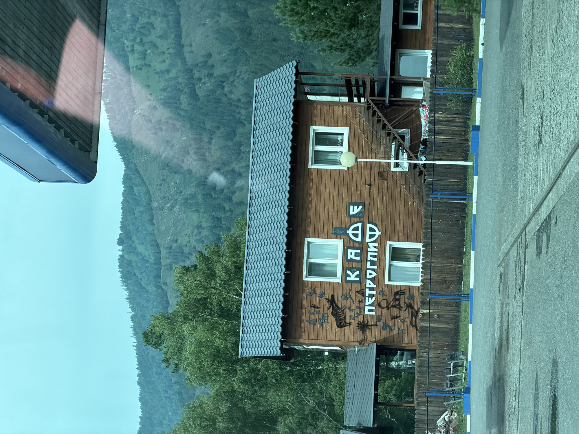 A wooden two-story building with a gray, wavy roof stands beside a wooden fence. The building has white-framed windows and an external staircase leading to the upper floor. Various petroglyph-like drawings are painted on the wooden facade. Text in black Cyrillic letters reads "КАФЕ ПЕТРОГЛИФ" above a lower window. Behind the building, a lush, tree-covered mountain slope rises under a cloudy sky. In front of the fence, a blue and white striped border runs along a paved area. The image appears to be taken from a vehicle, indicated by the reflection of a roof structure in the upper portion of the frame. The location is identified as Katun, Russia.