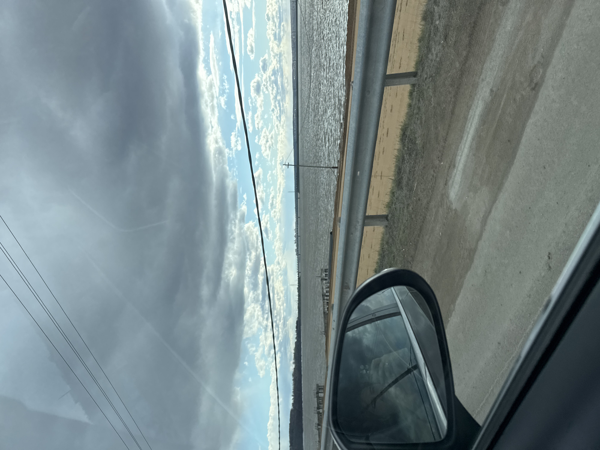The image captures a roadside view from a moving car, looking out over a vast body of water. In the lower right, the car's side-view mirror is prominent, reflecting the sky and possibly parts of the vehicle's interior.
The upper portion of the frame is dominated by a dynamic sky, with dark, heavy clouds on the left transitioning to brighter blue patches with fluffy white clouds towards the right, suggesting a mix of sun and cloud cover. Black power lines or utility wires stretch across the top left against the cloudy backdrop.
A wide expanse of water, possibly a large river or reservoir, fills the middle ground, its surface appearing textured and reflective, indicating light or a gentle ripple. Along the distant horizon across the water, a faint landmass or shoreline is visible, possibly dotted with distant structures or hills. Several small, dark, vertical posts or pilings are sparsely distributed in the water closer to the foreground.
Immediately to the right of the water, a grey metal guardrail runs horizontally, separating the road from the water. Beyond the guardrail, there's a narrow strip of dry, light brown ground with sparse patches of grass. A paved road surface is partially visible in the bottom right corner.
The scene depicts travel along a road that borders this large body of water, under partly cloudy skies during daytime. The setting suggests a semi-rural or an open area, consistent with the provided location context of Пальники, Russia, which is likely near a significant waterway or reservoir. No visible text is present in the image.