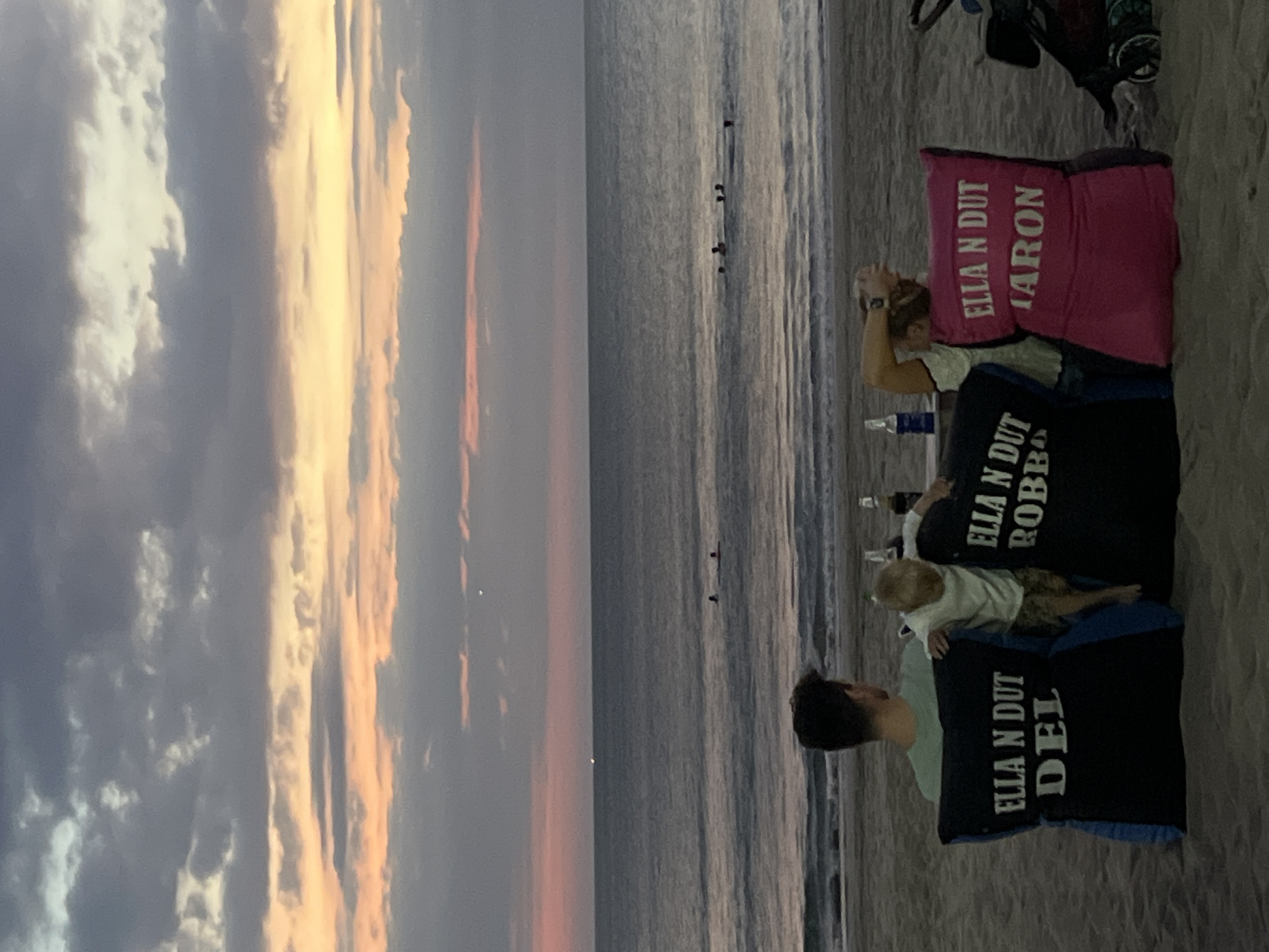 Captured at dusk in Kuta, Indonesia, the image presents three adults and one child seated on the sandy beach. Each person is reclining against a large, personalized cushion; the pink cushion is labeled "ELLA N DUT ARON", while the blue cushions bear the names "ELLA N DUT ROBBI" and "ELLA N DUT DEL." The adults are facing the ocean, where several people are seen standing or swimming. A partially visible beach cart with bags and equipment sits near the group. A few bottles are scattered on the sand. The sky is a blend of grays, yellows, and pinks, suggesting a sunset or sunrise.