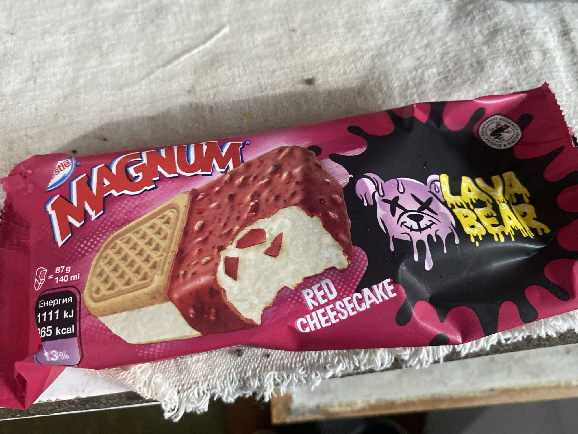 A single, partially crumpled, vibrant pink and black Magnum ice cream wrapper is the central subject, resting on a light-colored, woven textile surface. The wrapper displays an image of a 'Red Cheesecake' flavored ice cream bar, partially eaten to reveal a white, creamy interior, a waffle-textured biscuit layer, and a red, textured outer coating containing white chunks. The brand name 'MAGNUM' is prominently featured in red and white lettering, with the 'Nestlé' logo visible above it. The flavor 'RED CHEESECAKE' is printed vertically along the left side of the wrapper. Nutritional information on the top left indicates 'ЕНЕРГИЯ' (Energy) as 1111 kJ and 265 kcal per serving (87g = 140ml). A '13%' daily value percentage is also present. The bottom section of the wrapper features a stylized, pink cartoon character with an 'X' eye and a dripping mouth, set against a black background with pink splatters. Below the character, yellow text appears to read 'PLAYFUL'. A circular 'Rainforest Alliance' certification seal is visible on the bottom right corner. The background consists of a light beige or off-white fabric with a visible weave, possibly a towel or tablecloth. A fringed white edge and a darker, blurred surface are partially visible along the left side of the frame. The image does not contain any discernible information to specify its location within the city of Smolyan, Bulgaria, or any other specific geographic location.