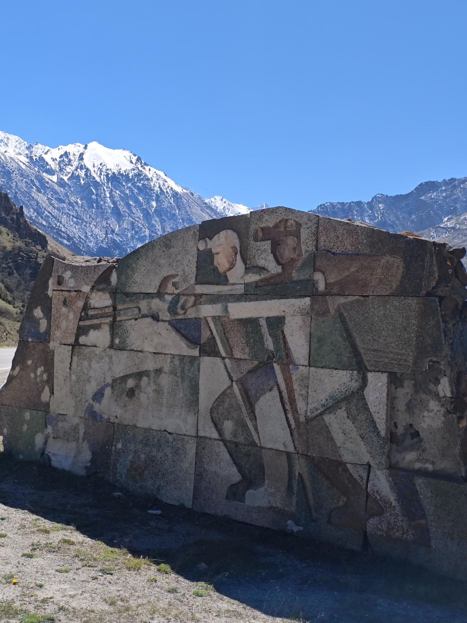 The image shows a monumental stone relief depicting two stylized figures, set against a backdrop of snow-capped mountains under a clear blue sky. The relief, constructed of large, irregular stones, features a lighter-skinned figure with a bare head and neck, and a darker-skinned figure wearing a hat. The figures are rendered in a geometric, angular style with visible brushstrokes and color variations. The relief is located on a gravelly, barren ground. This may be located in the vicinity of Tyrnyauz, Russia.