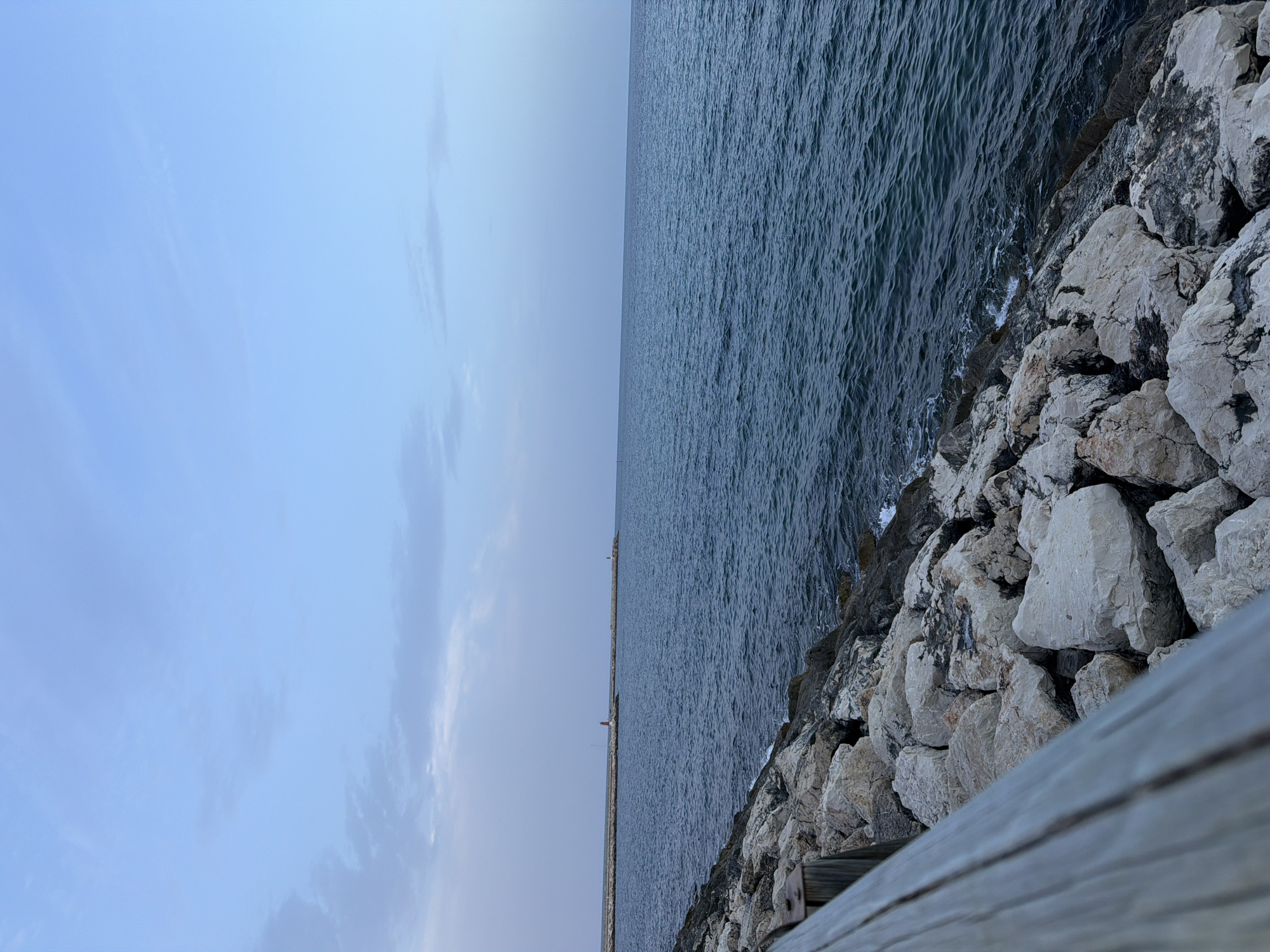A low-angle, slightly blurry shot captures a section of the rocky shoreline in Dénia, Spain, with the calm Mediterranean Sea to the right. A wooden barrier runs diagonally across the left foreground, obscuring a portion of the rocks. The rocks themselves are a mix of light and dark gray, with some appearing weathered and rounded, others more angular. Small waves gently break against the shore, creating white foam.
In the distance, two breakwaters extend into the sea. The closer breakwater, to the left of center, features a small red and white lighthouse. Further out, a second breakwater has a small green structure on its end. The sea is a deep, dark blue, reflecting the overcast sky. The sky itself is a pale blue with scattered, wispy clouds. The overall atmosphere is serene and tranquil. No people are visible in the frame.