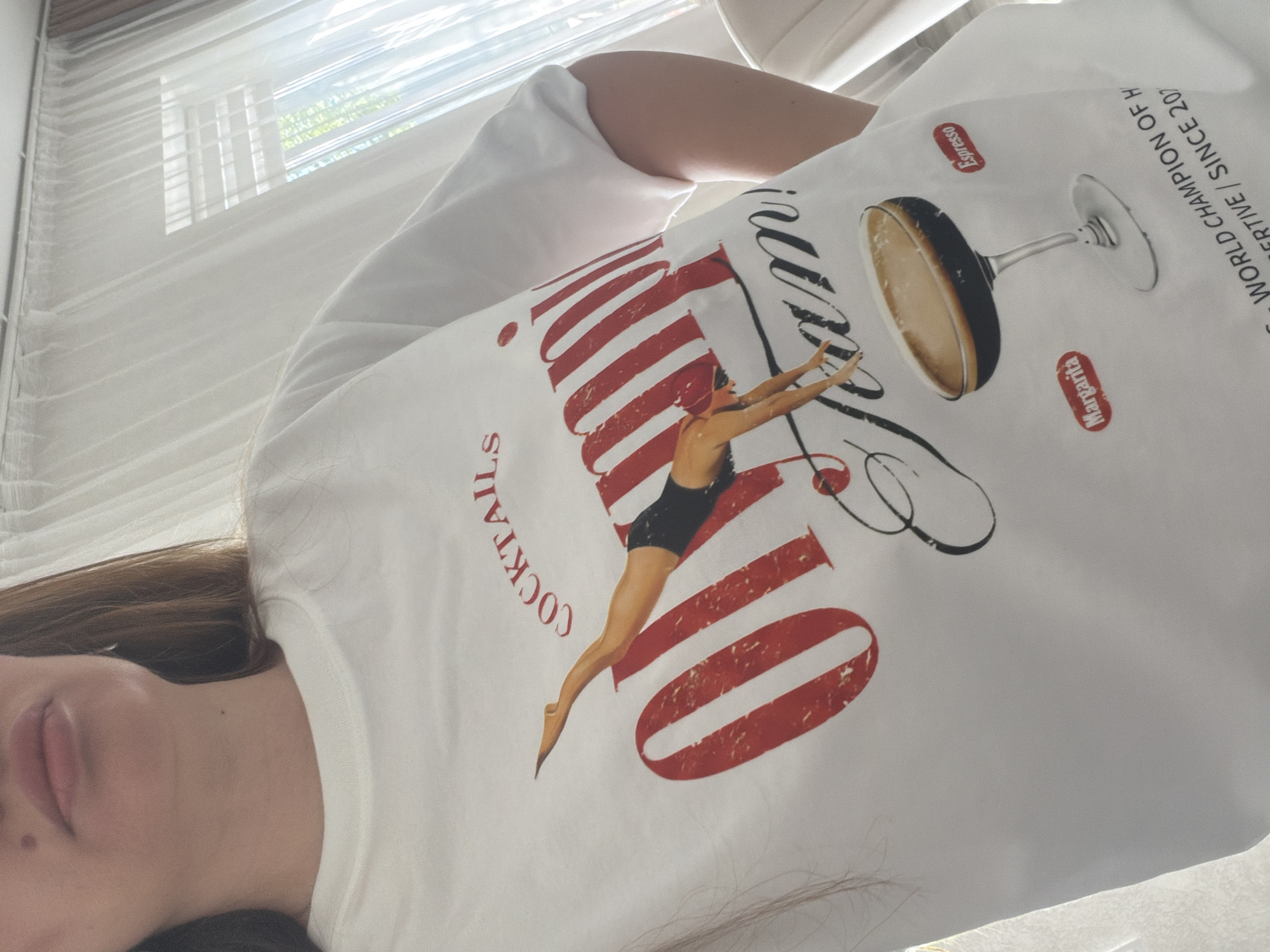 The image shows a white t-shirt with a graphic design. The graphic features a diving silhouette of a woman in a black swimsuit, positioned over large red text that reads "OLYMPIC TEAM!". To the left of the text, in red, are the words "COCKTAILS". Below the text and diver, there is a stylized drawing of a martini glass filled with a dark liquid, presumably espresso. To the left of the glass, in a red oval, is the word "MARGARITA", and to the right, in a red oval, is the word "ESPRESSO". At the bottom of the t-shirt, partially visible text reads "WORLD CHAMPION OF H...ERTIVE / SINCE 20...". The t-shirt is being worn by a person whose upper body is visible from the chest up. The background appears to be an indoor setting with sheer white curtains and natural light filtering through. The image does not contain any information or visual cues that would allow for its location to be identified as Malinovka, Russia.