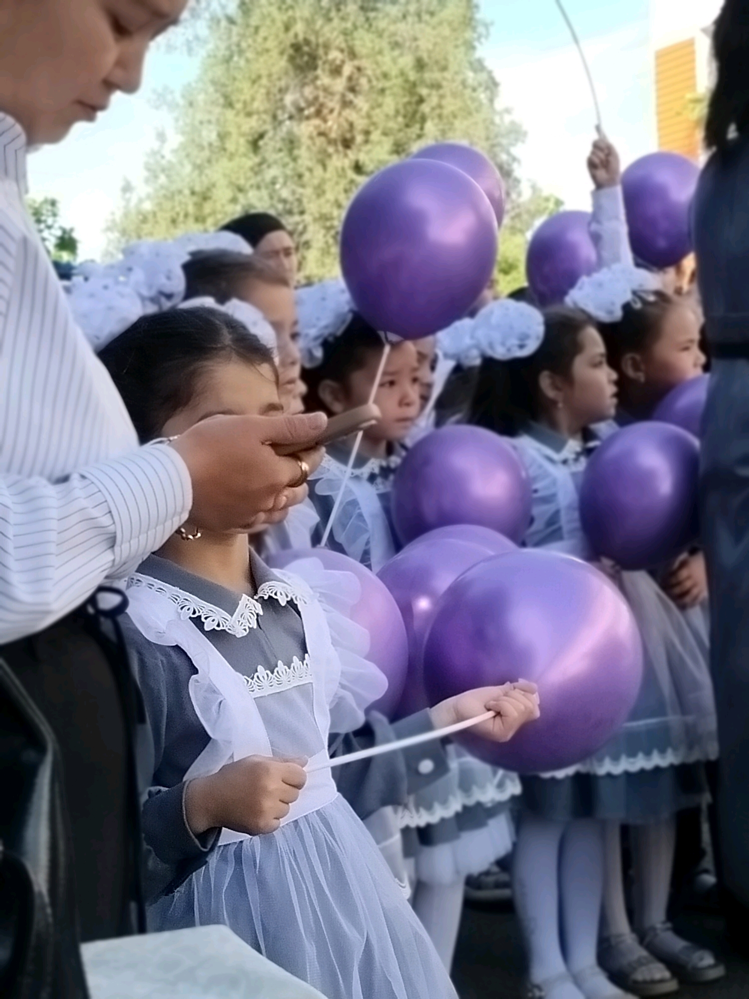 In the city of Kölkent, Kazakhstan, on what appears to be a sunny day, a group of young schoolgirls in uniform are gathered, holding purple balloons. The girls are wearing light blue dresses with white lace collars and aprons, and many have large white bows in their hair. One girl in the foreground is looking down, holding the string of a balloon, while an adult's hand with a ring is touching her face. Other girls are visible in the background, also holding balloons, some of which are partially obscured. The environment suggests an outdoor setting with trees and a building in the distance. The atmosphere seems festive, possibly marking the beginning of the school year.
