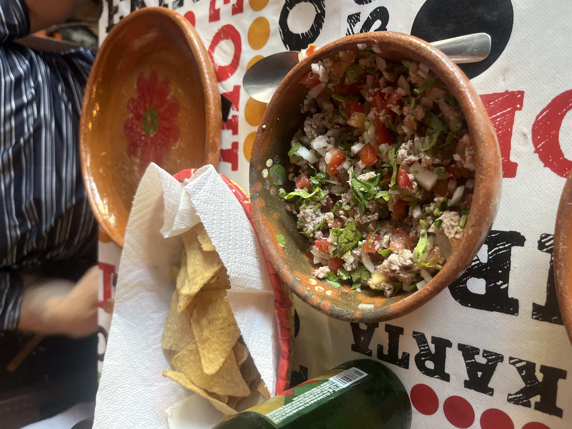 A table at a restaurant or home in San Francisco Totimehuacán, Mexico, is covered with a white tablecloth printed with black and red letters and circles. On the table are two earthenware bowls. The first bowl is empty and decorated with a red flower. The second bowl is filled with what appears to be a mixture of ground meat, tomatoes, onions, and cilantro, and a silver spoon rests inside. Next to the bowls is a red basket lined with white paper towels, containing a pile of tortilla chips. A green glass bottle is on the table near the basket. A person wearing a striped shirt is partially visible in the background.