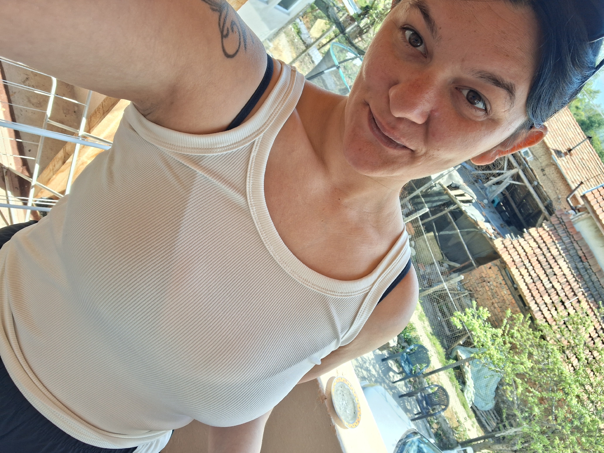 A woman is taking a selfie outdoors. She is fair-skinned with dark hair and eyes. She is wearing a light-colored tank top with black bra straps visible. A tattoo is visible on her left upper arm. The background shows a rural setting, potentially near the city of Gotse Delchev, Bulgaria. Elements in the background include a brick building with a tiled roof, a wire fence, trees with green foliage, and plastic chairs. The location appears to be a yard or garden area.