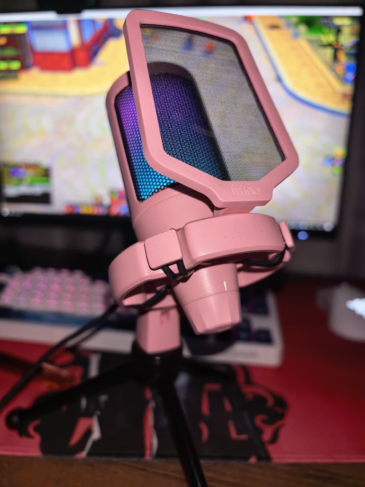 A close-up, low-angle shot shows a pink microphone with a pop filter positioned in front of a computer monitor displaying a colorful, pixelated game interface. The microphone has an LED light strip that transitions through purple and blue hues. It is mounted on a black tripod stand, which is placed on a red desk mat with a black graphic design. A pink-backlit keyboard and a white computer mouse are partially visible on the desk behind the microphone. The background is out of focus, with the computer monitor being the most discernible element. The lighting is dim, with the microphone's LED lights providing the primary illumination.