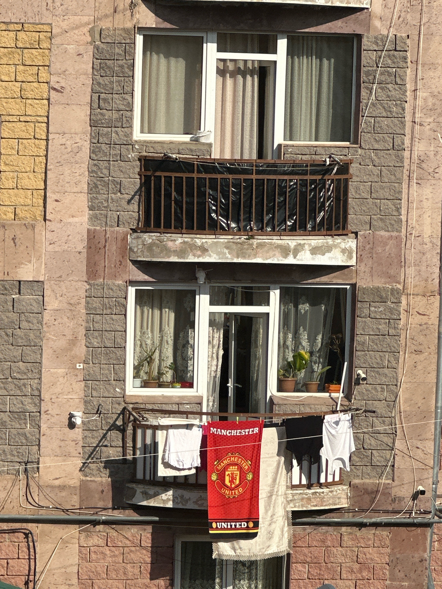 The image shows a residential building in Gyumri, Armenia, with balconies and windows on its facade. On one of the balconies, a Manchester United flag is hanging alongside laundry, including a white t-shirt and dark shorts. There are also two security cameras visible on the building's exterior. The building appears to be constructed with stone and brick. The lighting suggests it is daytime.