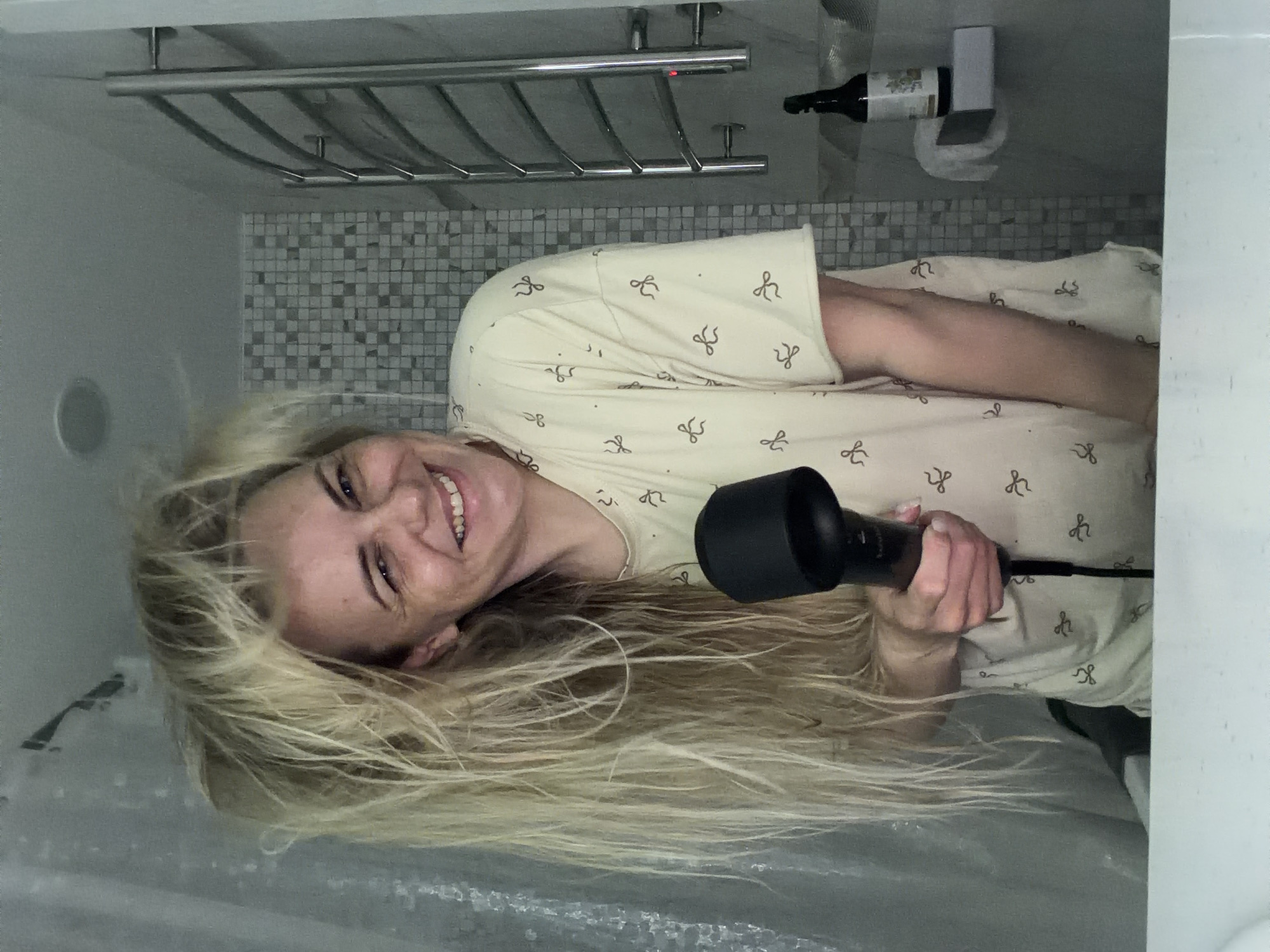 The image, rotated 90 degrees counter-clockwise, depicts a fair-skinned person with long, flowing blonde hair, captured mid-action. The person is smiling broadly and looking towards the camera, conveying a happy or joyful emotion. They are wearing a light-colored top or pajama shirt patterned with small, dark bow or ribbon shapes.
In their right hand, the person holds a black hairdryer fitted with a diffuser attachment, with their long hair spread out around them, indicating they are in the process of drying their hair. The setting appears to be an indoor space, likely a bathroom, given the background elements. These include a wall with light grey and white mosaic tiles, above which a metallic, multi-barred towel rack or shelf is mounted. To the upper right, a dark wine bottle with an unreadable label is secured in a white, wall-mounted dispenser or holder. The location context for this scene is Votkinsk, Russia.
