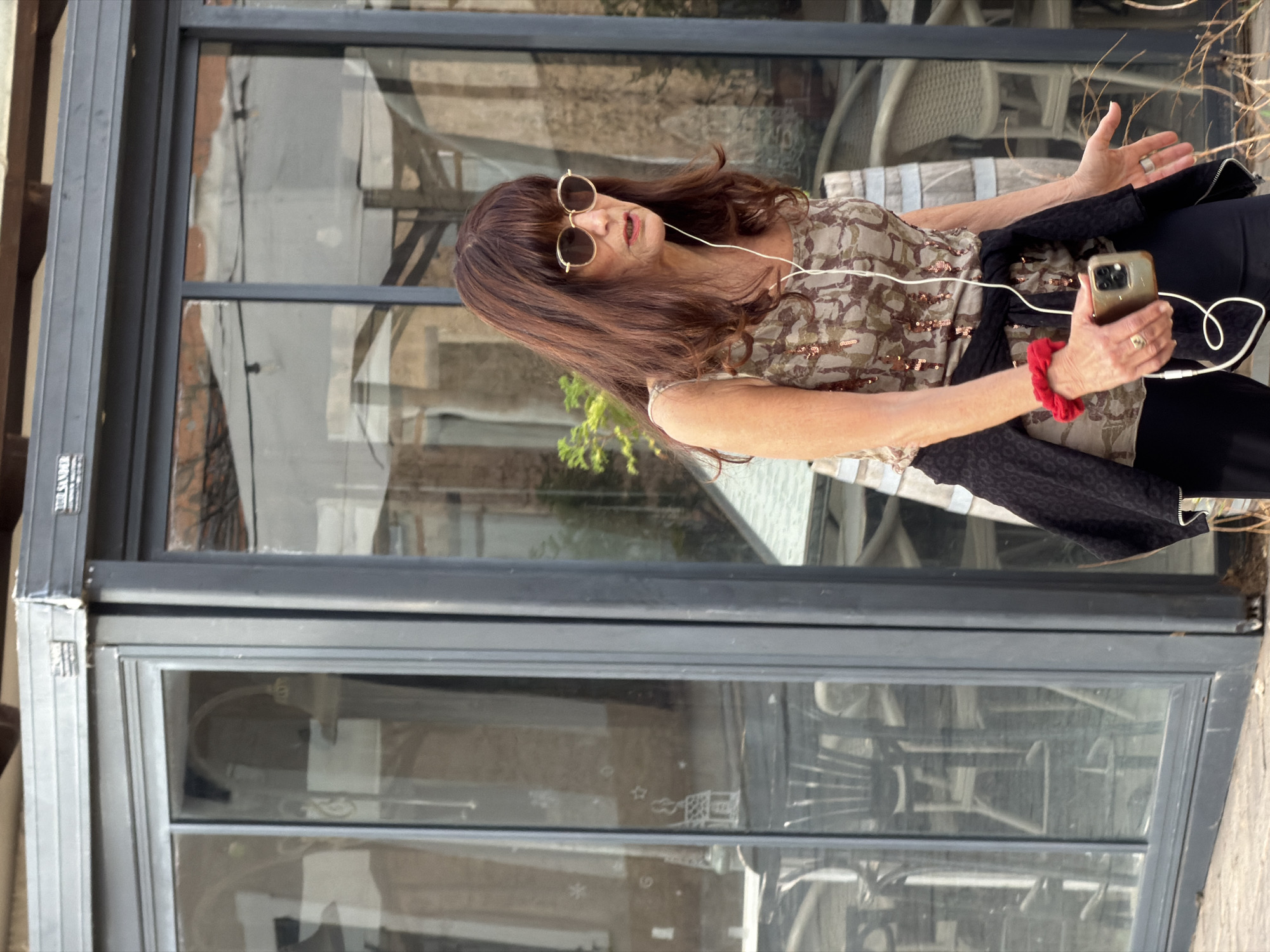 A middle-aged female is depicted in a full shot, walking outdoors in Tel Aviv, Israel, in front of a commercial establishment. She has long, dark brown hair with bangs and wears gold-rimmed round sunglasses. Her attire includes a sleeveless, sequined top with a camouflage-like pattern in beige and brown tones, black pants, and a black textured jacket tied around her waist. She wears white wired earphones, connected to a light-colored smartphone held in her right hand. A red scrunchie is on her left wrist, and multiple rings are visible on her fingers. Her mouth is slightly open, showing red lipstick.
The background features the facade of a building, likely a restaurant or cafe, characterized by large, dark-framed glass sliding doors and windows. A dark grey retractable awning structure is visible above the glass, displaying the word "TEL-AVIV" on a label, confirming the city. Reflections on the glass reveal internal seating arrangements with light-colored chairs and tables, as well as exterior elements. A partial wooden barrel is positioned to the right of the subject, and a small green plant is visible behind her left shoulder. The ground is a light-colored paved surface. The subject appears to be in motion, engaging with her phone and earphones.