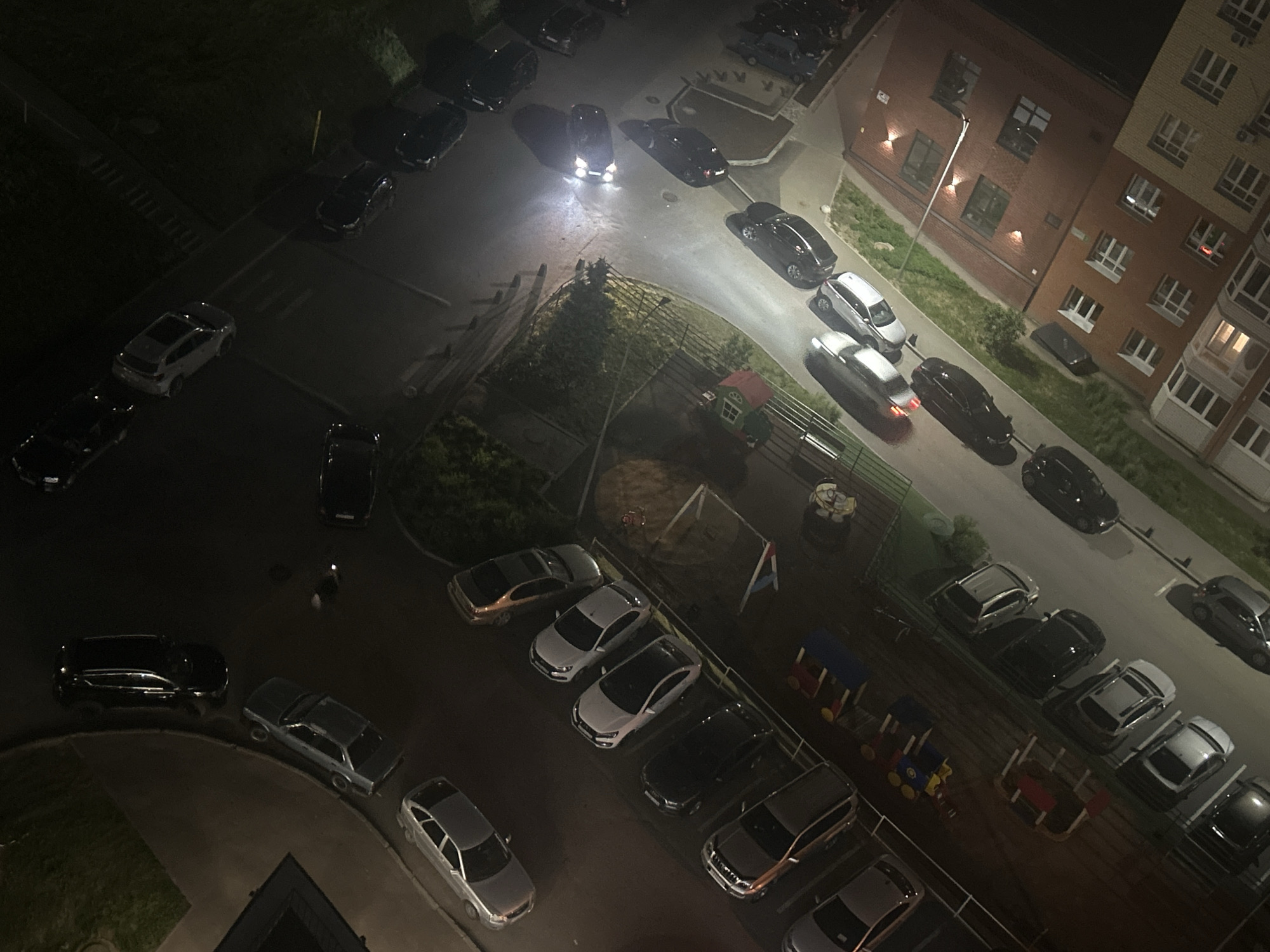 The image shows an aerial view of an outdoor area, likely a residential neighborhood in the city of Unknown, Unknown at night. There are multiple parked cars along streets and in parking lots. A building with lit windows is visible in the background. There appears to be a playground with a slide and some toy structures. A person can be seen walking in the lower left quadrant of the image. The lighting is dim with some areas illuminated by headlights and streetlights.