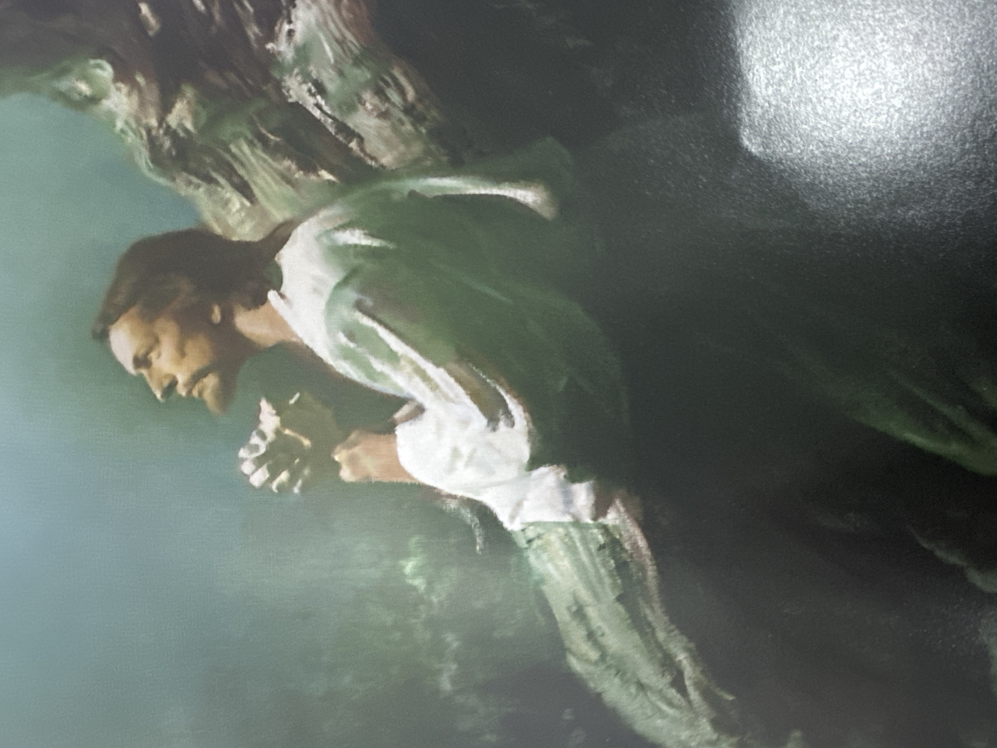 The image is a painting or digital artwork featuring a solitary man in a dark, natural environment. The man, depicted in profile, is positioned on the left side of the frame, facing towards the right and looking downwards. He has short, dark hair and a beard, and is dressed in a light-colored, long-sleeved tunic or shirt and darker, possibly green or brown, trousers. His hands are clasped together in front of him, suggesting a posture of prayer, supplication, or deep contemplation. His expression appears solemn and focused.
The background is predominantly dark and indistinct, featuring what appears to be a rocky or uneven natural formation above and behind the man, with subtle hints of green, possibly foliage or moss. A soft, diffused light emanates from the upper left, illuminating parts of the man's face, clothing, and the immediate surroundings. The overall lighting is dim, creating a serious and introspective mood, possibly suggesting a night scene or a deeply shaded location. The artwork displays a painterly quality with visible textures. No text is visible in the image. While "Jos, Nigeria" is provided as a context, no specific visual elements within the image directly identify this location.