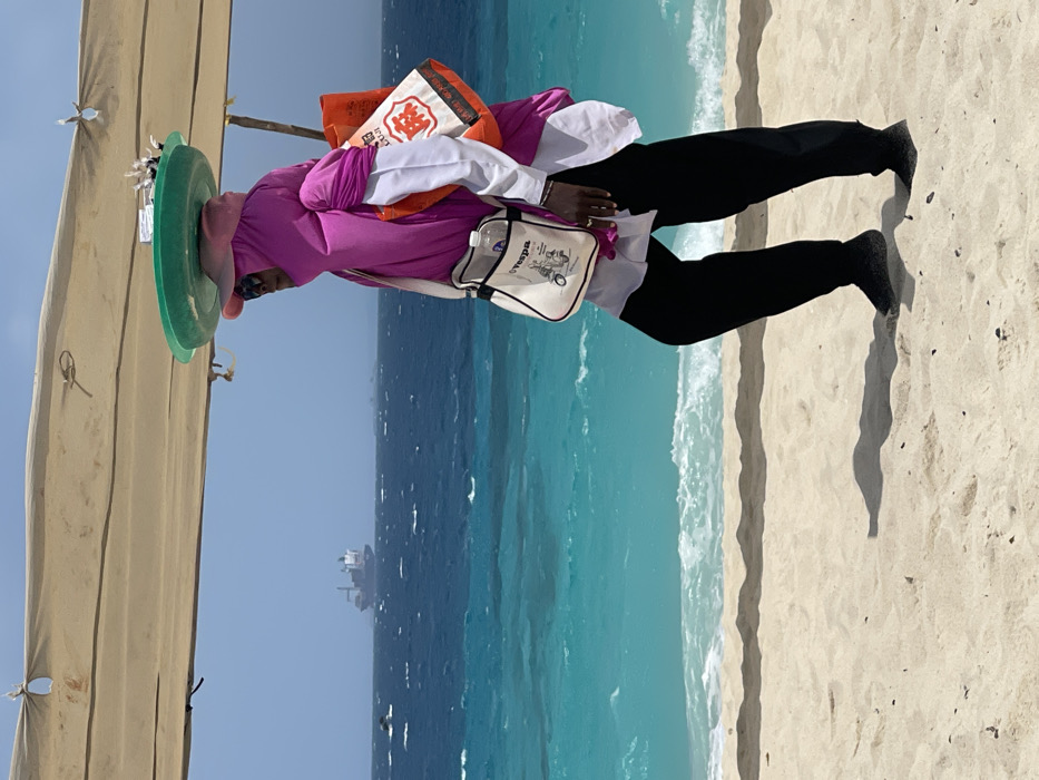 An individual walks rightward on a light sandy beach in Zanzibar, Tanzania, beneath a partial beige fabric canopy. The person wears a magenta full-body covering over a white long-sleeved shirt, black pants, and dark footwear. A pink baseball cap and sunglasses are visible beneath the head covering. A green, shallow, round tray holding small, dark objects is balanced on their head. They carry a white canvas shoulder bag with a "Vespa" graphic, containing a clear plastic water bottle, and an orange fabric tote bag with white Chinese characters, slung over their right shoulder. The person casts a distinct shadow on the sand. To the left, the turquoise ocean meets the shore with gentle white waves, transitioning to a deeper blue further from the coast. A large cargo ship is visible on the distant horizon against a light blue sky, with several smaller dark objects present in the mid-distance water.