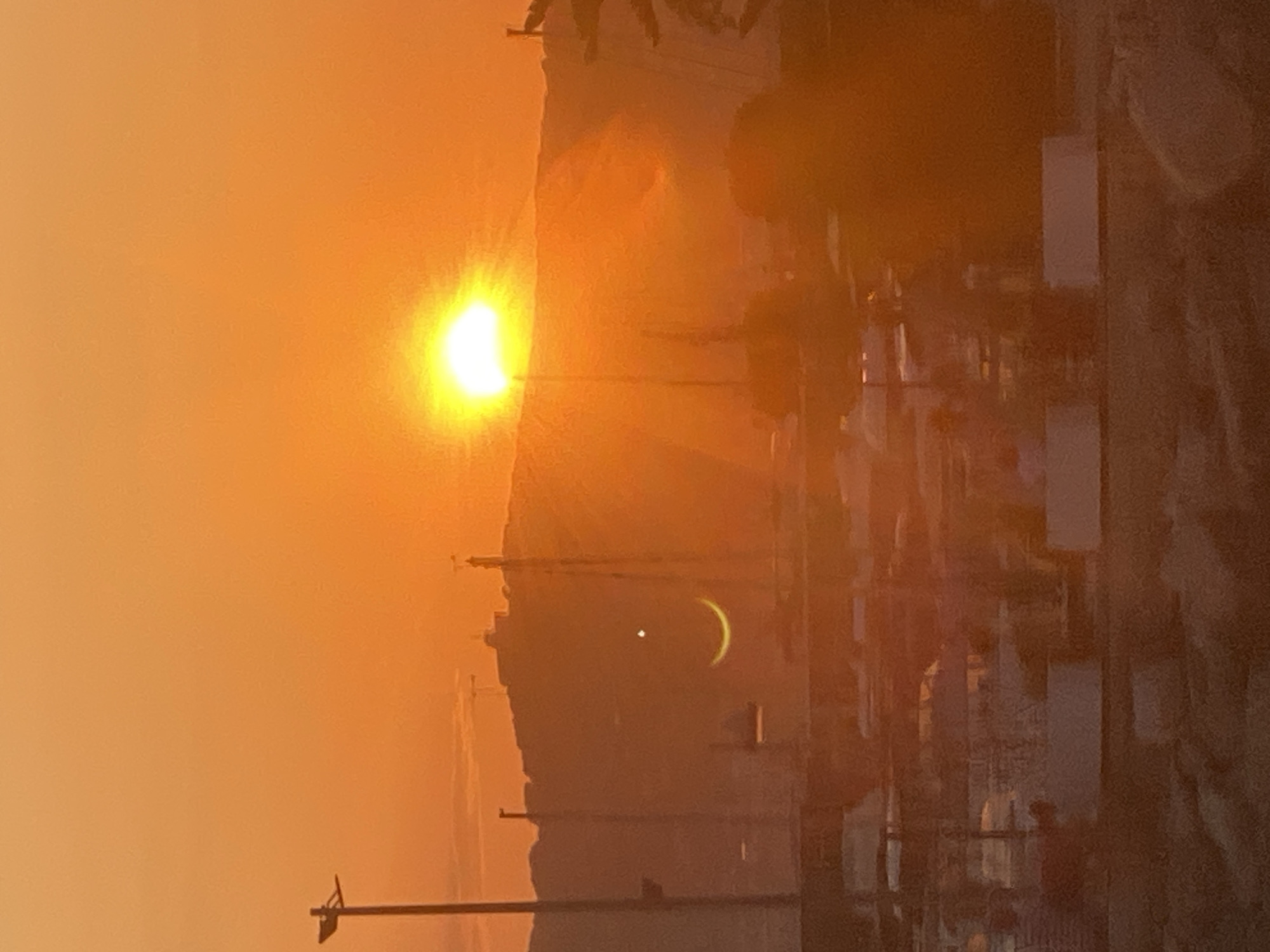 The image is a vertical shot of a sunset over a coastal city, possibly Antibes, France, given the context. The sun, a brilliant orb of light, is partially obscured by a hazy sky, casting a warm orange glow over the entire scene. In the foreground, silhouetted against the bright sky, are several utility poles and wires, suggesting an urban or semi-urban environment.
Further into the scene, the outline of buildings and structures is visible, their details softened by the atmospheric haze and the sunset's light. A small, crescent-shaped object, possibly the moon or a reflection, is visible in the sky, accompanied by a tiny, bright dot, perhaps a star or a planet. The overall mood is serene and tranquil, characteristic of a sunset. The colors are dominated by warm oranges and yellows, with darker silhouettes creating depth. There is no visible text in the image.