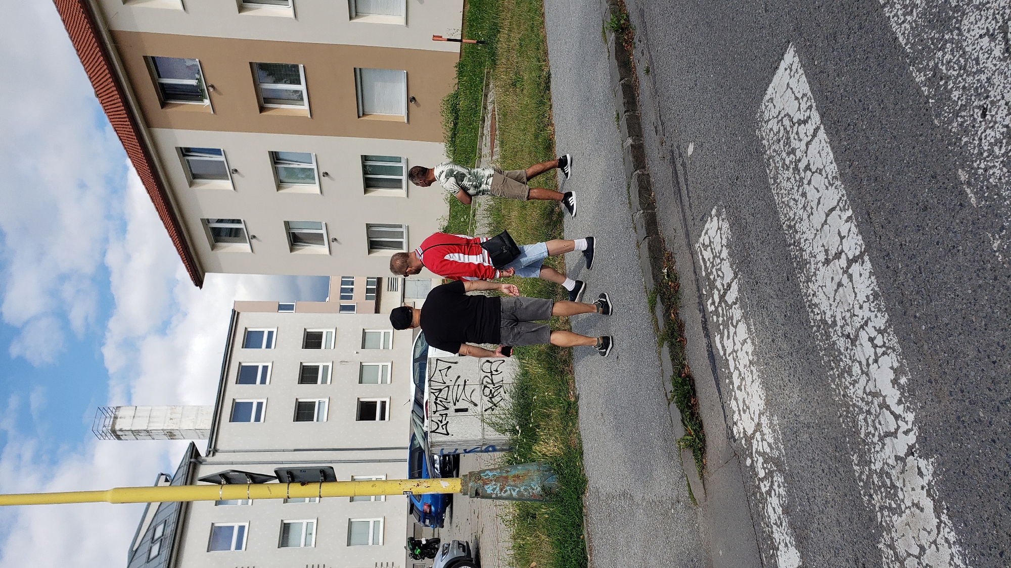 Three men are walking on a sidewalk next to apartment buildings in Prešov, Slovakia. The man on the left wears a black t-shirt and gray shorts. The middle man wears a red and white jacket and blue shorts, with a black bag slung over his shoulder. The man on the right wears a white, green, and brown patterned t-shirt and beige shorts. A yellow traffic pole with a sign is to their left, and a concrete structure with graffiti is behind the man on the left. The buildings appear to be residential, with multiple windows. A pedestrian crossing is visible in the foreground on the asphalt road. The sky is partly cloudy.