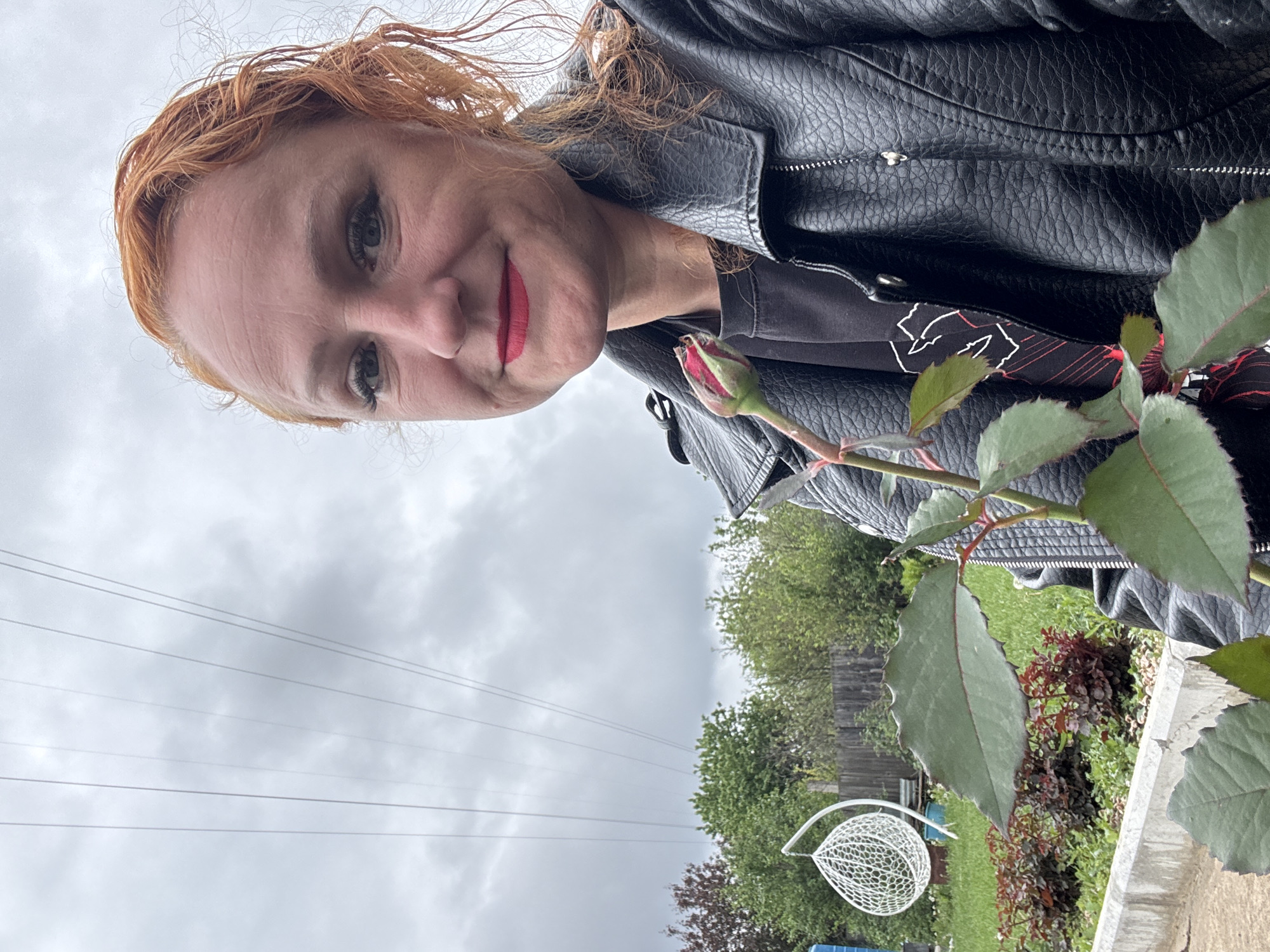 A selfie taken at an angle shows a woman with auburn hair, wearing a black leather jacket and a black t-shirt with a white design. She is holding a rose bud. The woman is wearing red lipstick.
In the background, a cloudy sky is visible with power lines. Below the power lines are trees and a wooden fence. A white, decorative wire structure is present near what appears to be a small garden area with different types of plants.
The location is potentially Chaikovsky, Russia, based on the request.