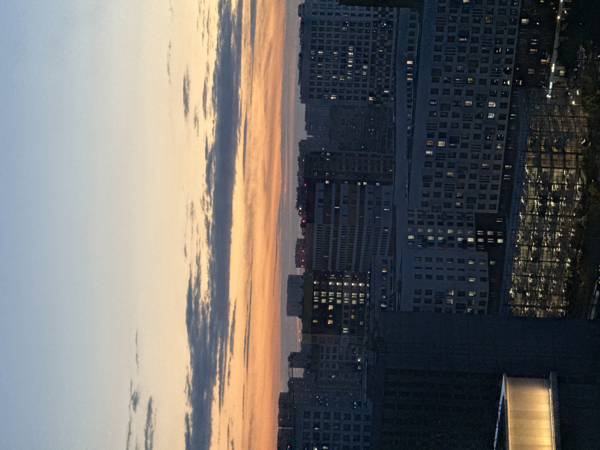 This image depicts a city skyline at dusk in Dolgoprudny, Russia. The sky transitions from a pale blue at the top to a soft orange and then darker gray clouds closer to the horizon. Several multi-story residential buildings, characterized by rows of lit windows, dominate the midground and foreground. One building in the foreground on the left features a brightly lit, curved section. Below the buildings, a large, glass-enclosed structure, possibly a parking garage, is visible with numerous lights illuminating its interior and many parked vehicles. Trees and some darker, less defined structures are present at the very bottom. The scene is characterized by the artificial lights of the buildings and the fading natural light of the sky.