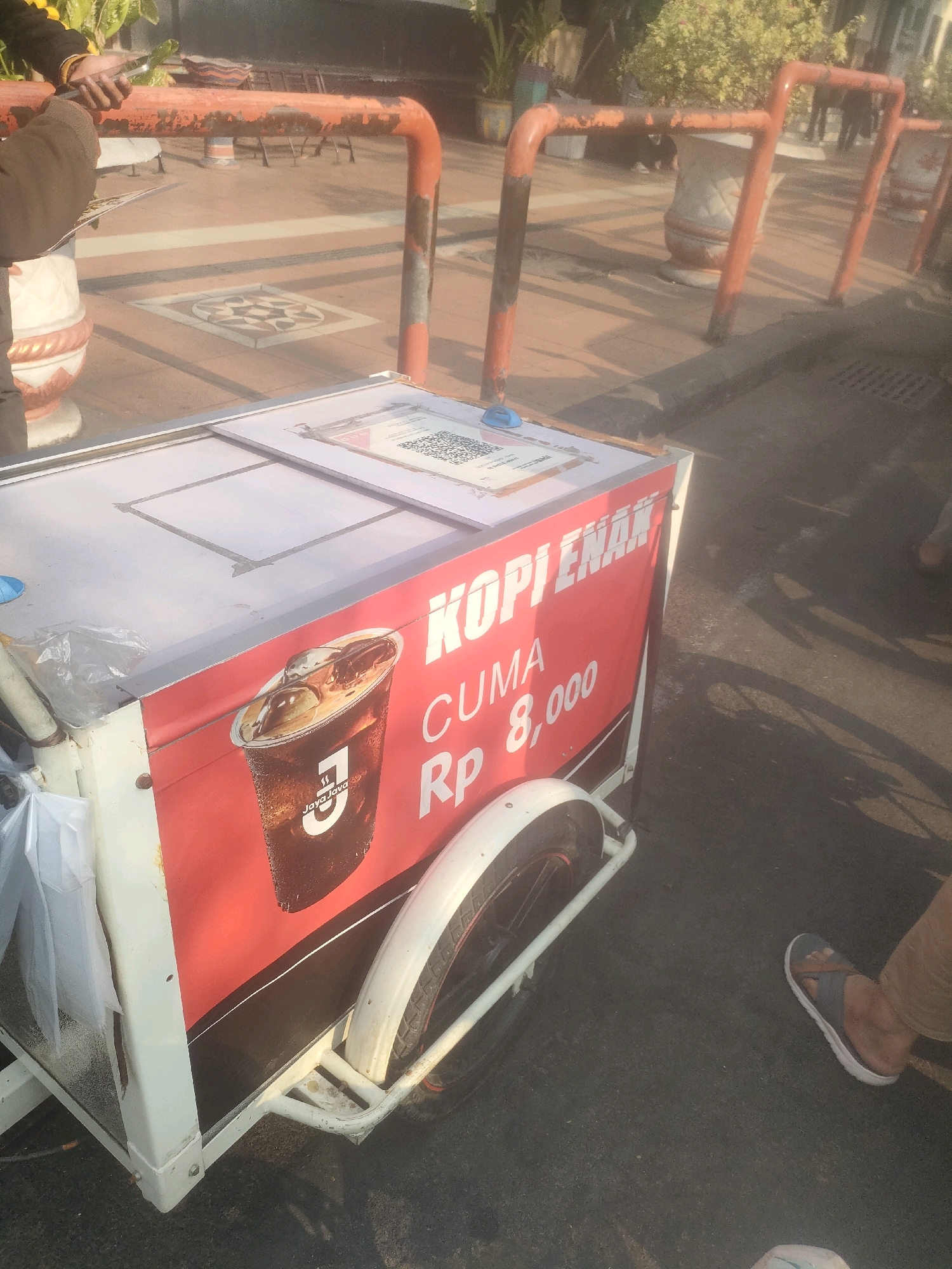 In Surabaya, Indonesia, on a paved outdoor area, a white wheeled cart is positioned. The cart has a red side panel featuring text and an image. The text reads "KOPI ENAK CUMA Rp 8,000" which translates to "DELICIOUS COFFEE ONLY Rp 8,000". An image of a large iced coffee drink is displayed on the red panel. The top of the cart has a white lid with a QR code sticker. A person's legs and feet are visible on the right side of the image, one wearing a sandal. The background shows an outdoor setting with trees and potted plants, suggesting a public space. The cart appears to be a mobile coffee vendor.