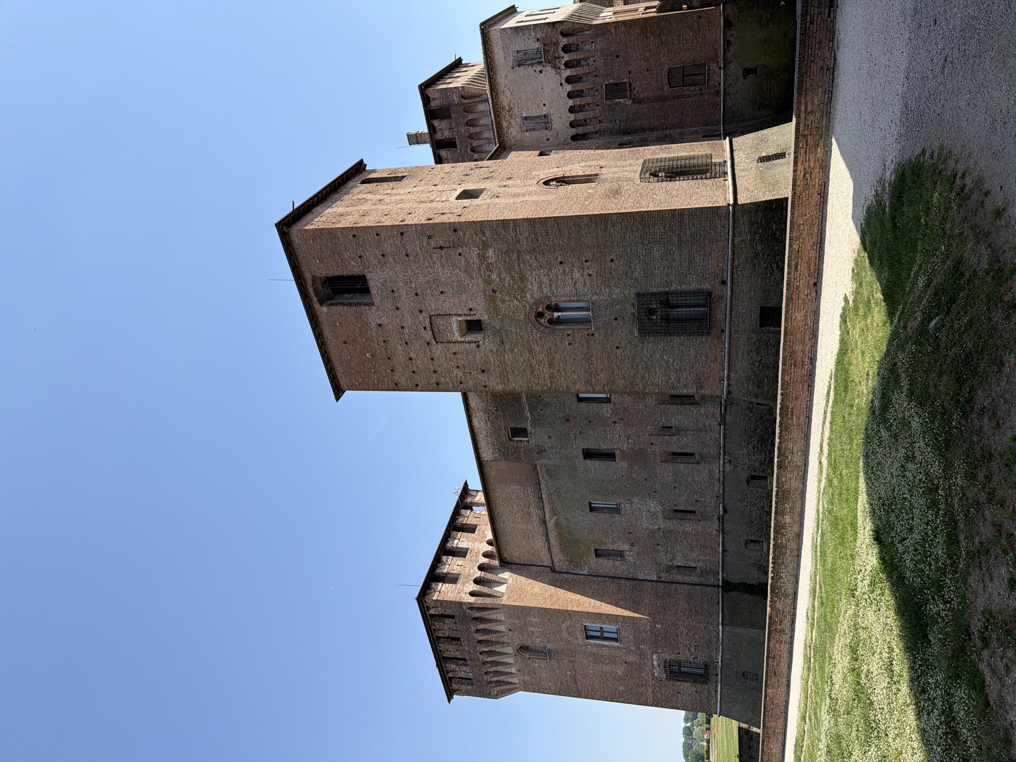 The image shows the Castello di San Giorgio in Mantua, Italy, viewed from a low angle looking up at the exterior brick facade and towers against a clear blue sky. The architecture is medieval, with crenellations visible on the upper parts of the towers. Below is a gravel path with green lawn in view on the side of the building. There are no people visible in the scene.