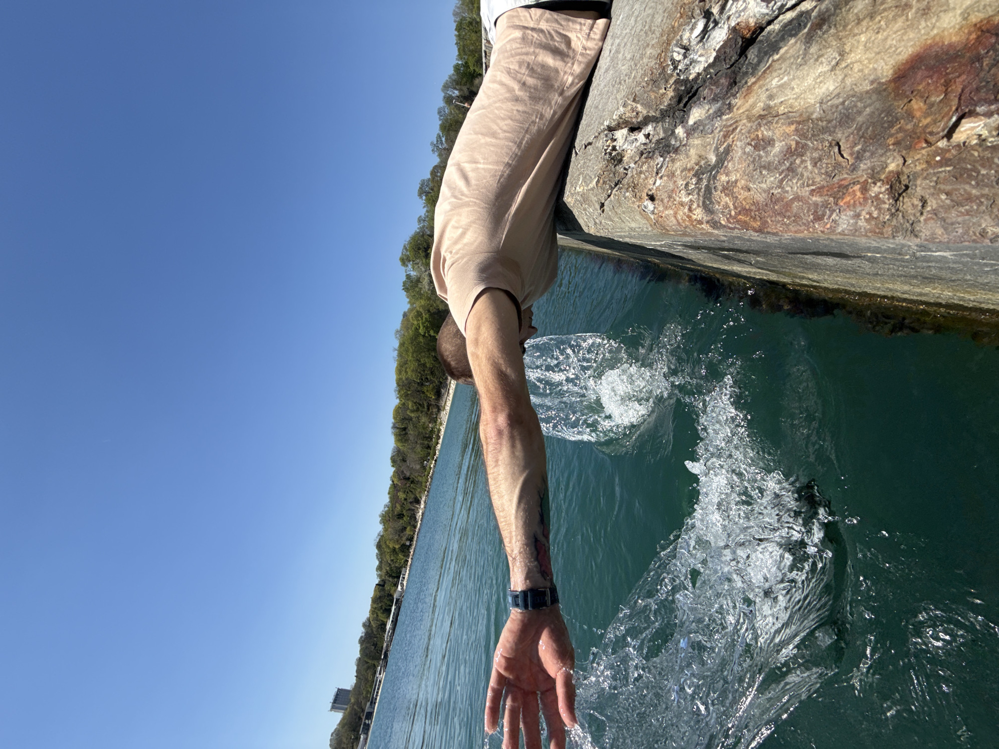 A man leans over a weathered stone pier, dipping his hand into the green seawater. Splashes erupt around his hand as it enters the water. He wears a light beige T-shirt and a blue wristwatch. The pier's edge shows signs of age and wear. Behind the pier, the coastline of Varna, Bulgaria, features trees and buildings along the shore. The sky is a clear, bright blue.
