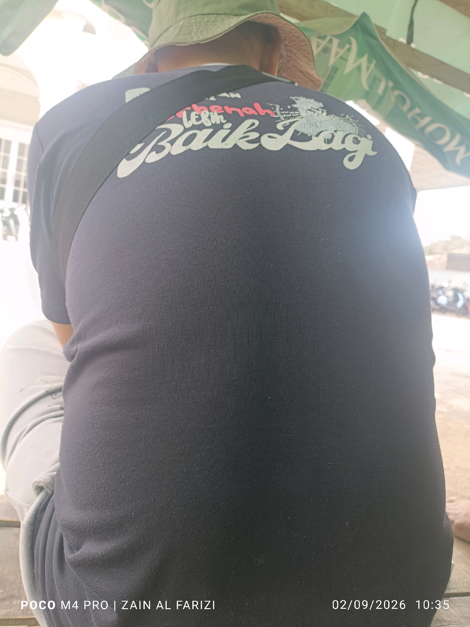 The image shows a person from the back, wearing a dark blue t-shirt with a graphic design on it. The design features white text with pink accents that reads "Lebih Baik Lagi." The person is also wearing light-colored pants and a hat. In the background, there are green banners with text that appears to be in Indonesian. The scene is outdoors, under a canopy. The lower right corner of the image contains a timestamp and text that reads "POCO M4 PRO | ZAIN AL FARIZI" and "02/09/2026 10:35". The image is likely taken in Cendagah, Indonesia, given the presence of Indonesian text and the recognizable context of a street scene.