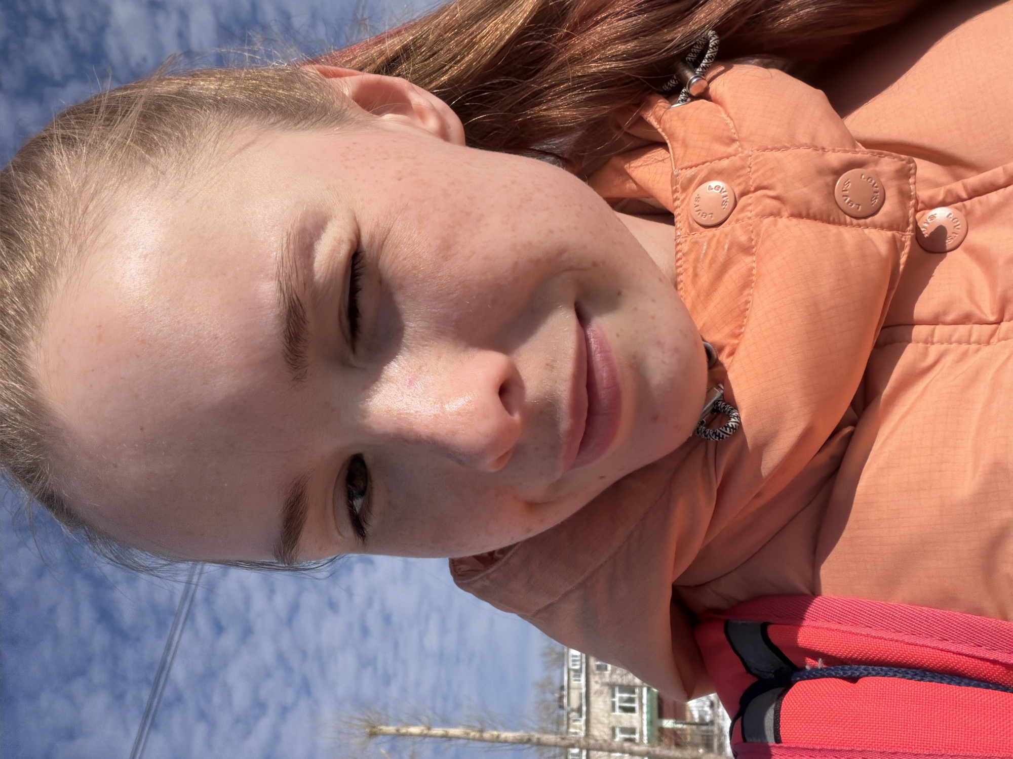 The image is a close-up portrait of a young person, rotated ninety degrees to the right. The subject, likely female, is captured from a low angle. She has light brown hair and fair skin with numerous freckles across her nose and cheeks. Her left eye is visible, while her right eye is partially closed or squinting due to the bright sunlight. A faint smile plays on her lips, suggesting a pleasant and casual mood.
She is wearing an orange or peach-colored puffer jacket with a textured surface. Several light-colored buttons are visible on the jacket's collar area, each embossed with the text "LOVLS". A black and white patterned drawstring, part of the jacket's design, is also visible near her neck. A bright red or pink backpack or bag is partially visible at the bottom right of the frame.
The background features a bright blue sky filled with scattered, wispy white clouds, indicating clear and sunny weather. Below the horizon, bare trees, characteristic of late winter or early spring, are visible. In the distance, indistinct multi-story buildings suggest an urban or suburban setting. The strong lighting and shadows imply it is daytime, likely midday or early afternoon. The location is Berezniki, Russia.
